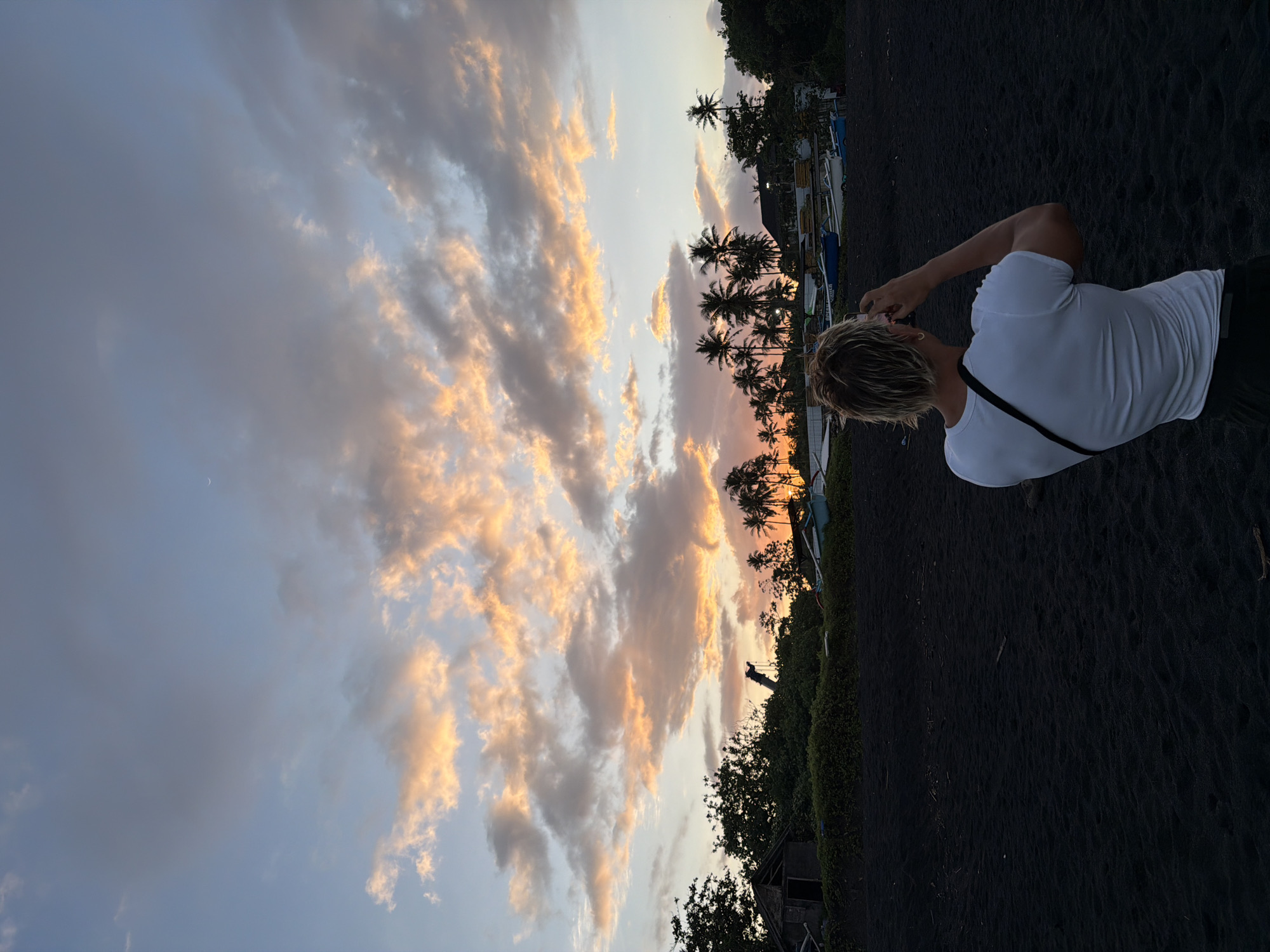 An eye-level, landscape-oriented photograph taken in Mengwi, Indonesia, shows a beach scene at sunset. A person wearing a white t-shirt and a black shoulder bag stands on black sand, their back to the camera, facing toward a line of silhouetted palm trees and low vegetation. The sky is filled with clouds tinted with orange and pink from the setting sun. The sand appears dark and textured. In the background, there are boats.