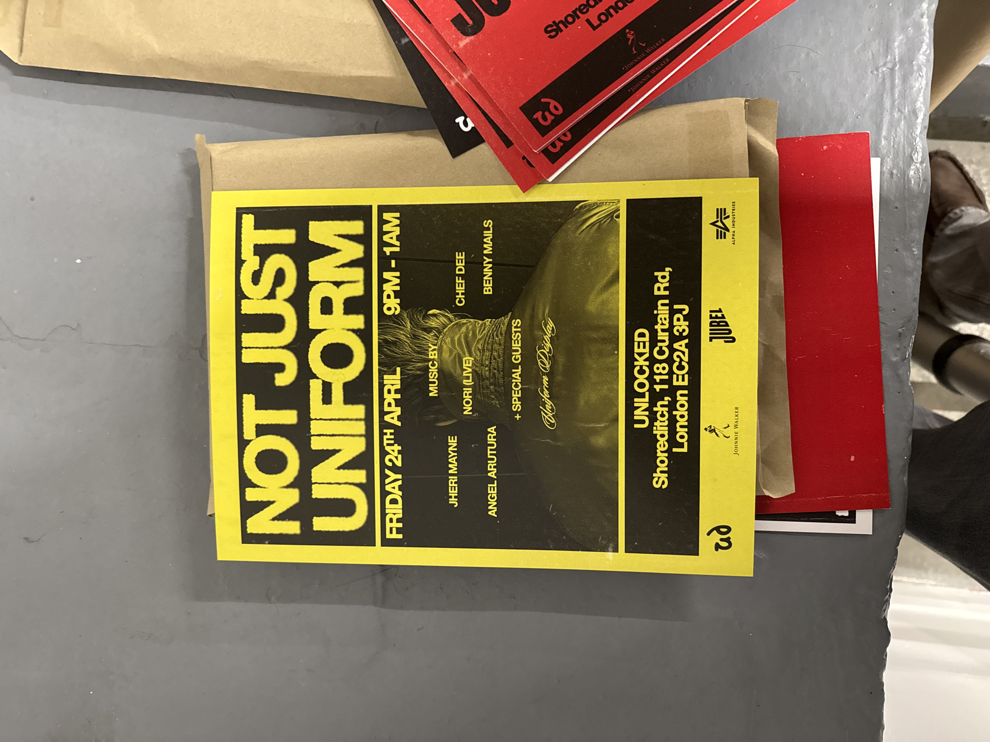 The image features a bright yellow event flyer laid on a grey, possibly concrete or metal, surface. The flyer is the central focus, surrounded by other paper items, including two brown paper envelopes or packages and several red cards. A partial view of a person's brown shoe and lower leg is visible in the background on the right.
The yellow flyer advertises an event titled "NOT JUST UNIFORM". Below the title, the date is stated as "FRIDAY 24TH APRIL" and the time as "9PM - 1AM". The "MUSIC BY" section lists "JHERI MAYNE", "NORI (LIVE)", "ANGEL ARUTURA", "CHEF DEE", and "BENNY MAILS". It also mentions "+ SPECIAL GUESTS" and "Uniform Display". The venue is "UNLOCKED", located at "Shoreditch, 118 Curtain Rd, London EC2A 3PJ". Several brand logos are visible, including "JUBEL", "ALPHA INDUSTRIES", and "JOHNNIE WALKER" (with its striding man logo). A stylized 'ud' or 'pn' logo is repeated on the flyer.
The red cards in the background, also bearing a similar 'ud' or 'pn' logo, show partial text like "Shoreditch, London" and "JOHNNIE WALKER", suggesting they are related promotional materials for the same or a similar event. The scene depicts a collection of promotional items for an event taking place in London.