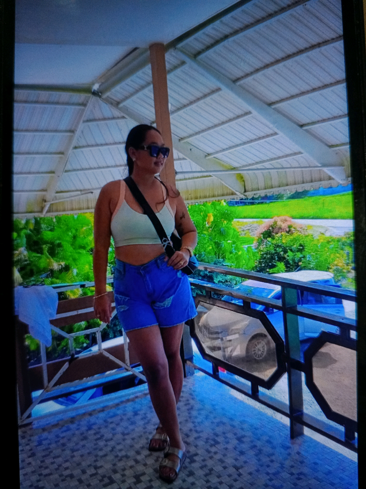 A full-body shot shows a woman standing on a tiled patio in Noord, Suriname. She is wearing blue denim shorts, a pale yellow crop top, and a black crossbody bag. She also has on dark sunglasses and gold sandals. The woman is looking to her right, with her left arm bent at the elbow and her right arm at her side. Behind her, there is a railing and a view of lush green trees and a bright blue car. The patio is covered by a white roof supported by wooden beams. The overall scene suggests a casual outdoor setting in a tropical location.