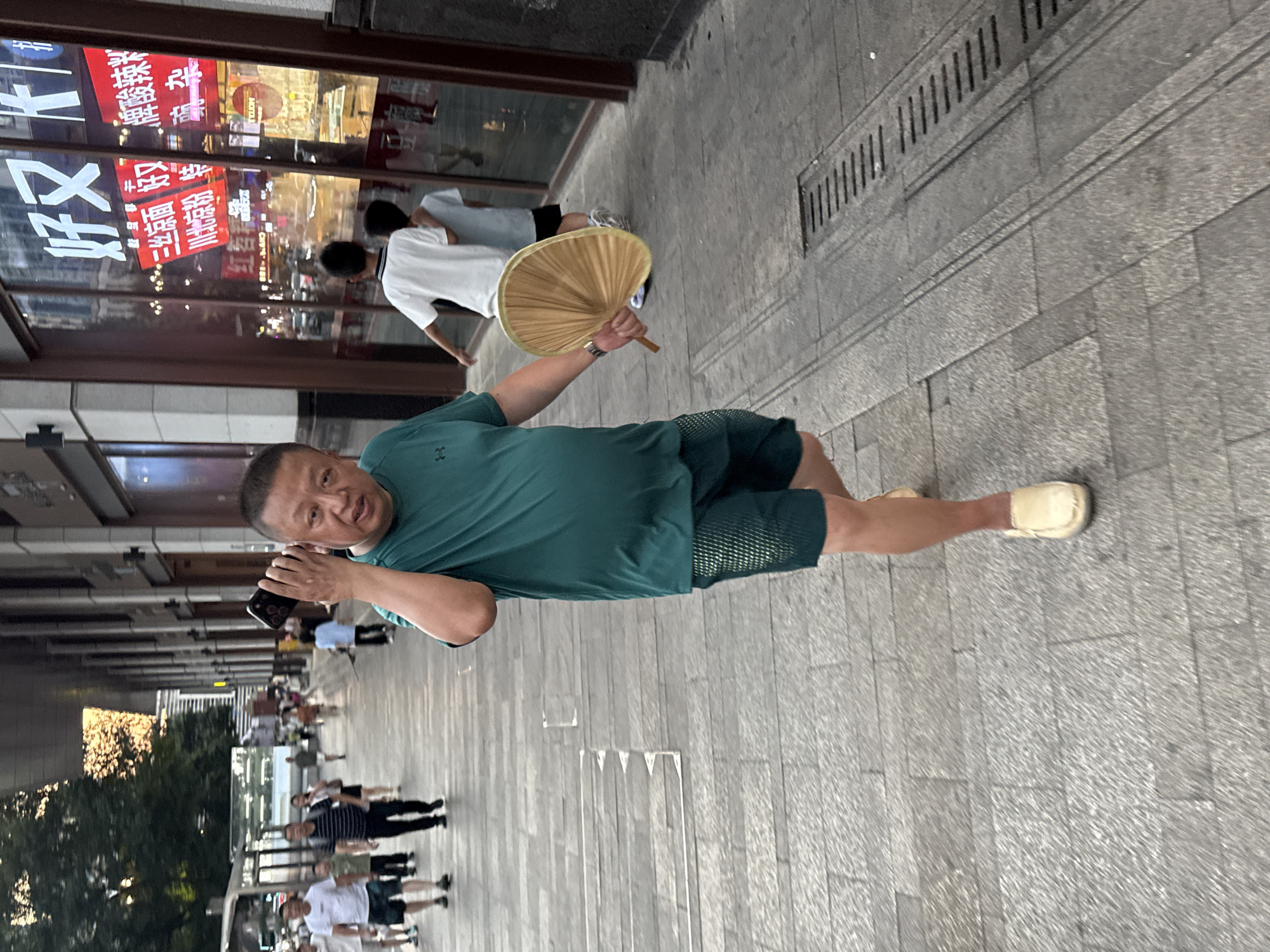 A medium shot captures a man in a green t-shirt and dark shorts walking on a paved sidewalk in Yuzhong District, China. The man, appearing to be in his 50s, holds a cell phone to his right ear with his right hand and a large, light-brown bamboo fan in his left hand. He is looking towards the camera with a neutral expression. Behind him, other pedestrians are visible on the sidewalk, some walking in the opposite direction. The background features the exterior of a building with large glass windows displaying Chinese text and illuminated signs. The sidewalk is made of gray stone tiles with a visible drainage grate. The lighting suggests it is daytime, possibly late afternoon, with some of the trees on the left side of the image bathed in sunlight.