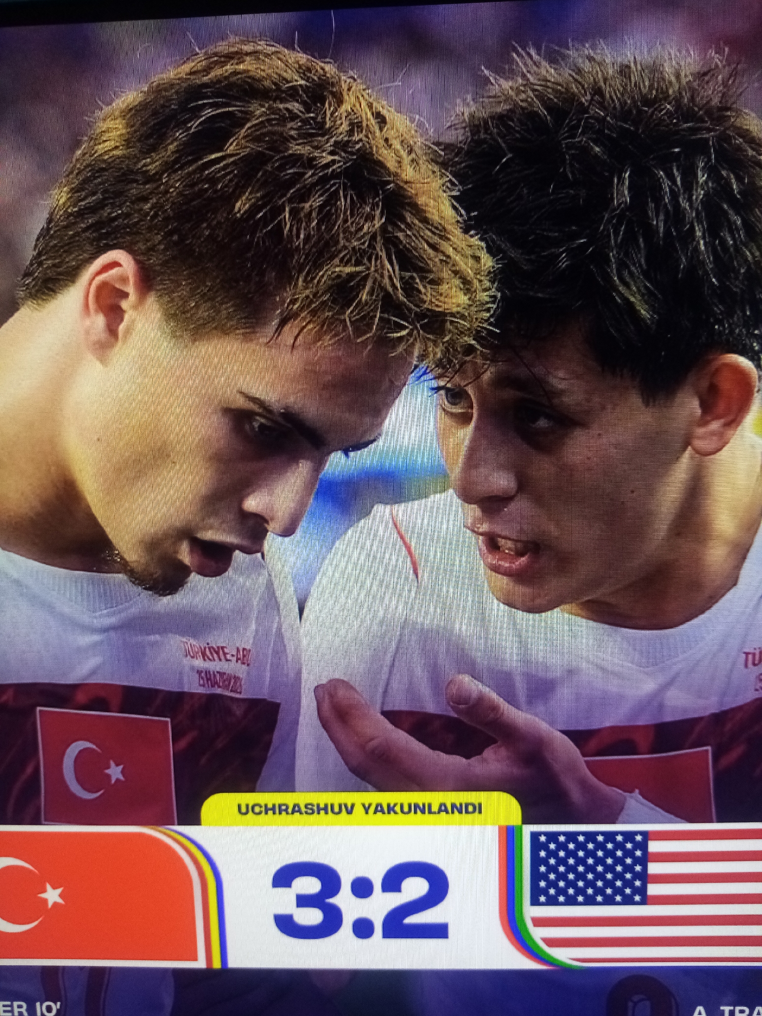 This media file captures a close-up view of two young men, likely athletes, engaged in an intense conversation. They are wearing white shirts with small flags and text indicating "TURKIYE-AR" which suggests they represent Turkey in a competition. The man on the left has light brown, spiky hair and is looking down, his mouth slightly open. The man on the right has darker, also spiky hair and is looking intensely at the other man, his mouth also open as if speaking. The background is blurred, but hints of purple and blue suggest an outdoor setting, possibly at dusk or under artificial lighting.
Below the men, a scoreboard-like display is visible. It features the Turkish flag, the text "UCHRASHUV YAKUNLANDI" (which translates from Uzbek to "The match has ended"), and the score "3:2". Next to the score is the American flag. This indicates that the event being depicted is likely a sports match between Turkey and the USA, with Turkey winning 3-2. The "ER 10'" text likely refers to the 10th minute of extra time or a similar game-related detail. The overall emotion conveyed is one of seriousness and possibly disappointment or urgency, given the intensity of their expressions and the context of a concluded match. The presence of flags and a score strongly places the scene within a sporting event.