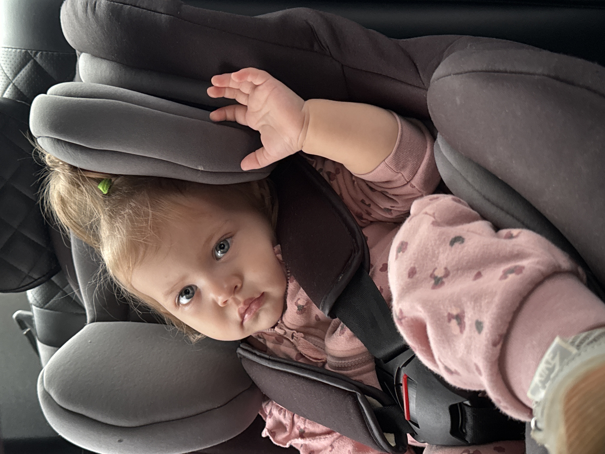 A close-up, high-angle shot shows a young child secured in a car seat. The child, with fair hair and wide blue eyes, is looking directly at the camera. They are wearing a pink outfit with a heart pattern. The car seat is dark gray and appears to be padded for comfort. The child's right hand is raised, resting on the side of the car seat. The background is dark and indistinct, suggesting the interior of a car. The lighting is even, indicating it is likely daytime. There is no visible text in the image. The overall impression is a candid moment of a child in a car seat, possibly on a journey.