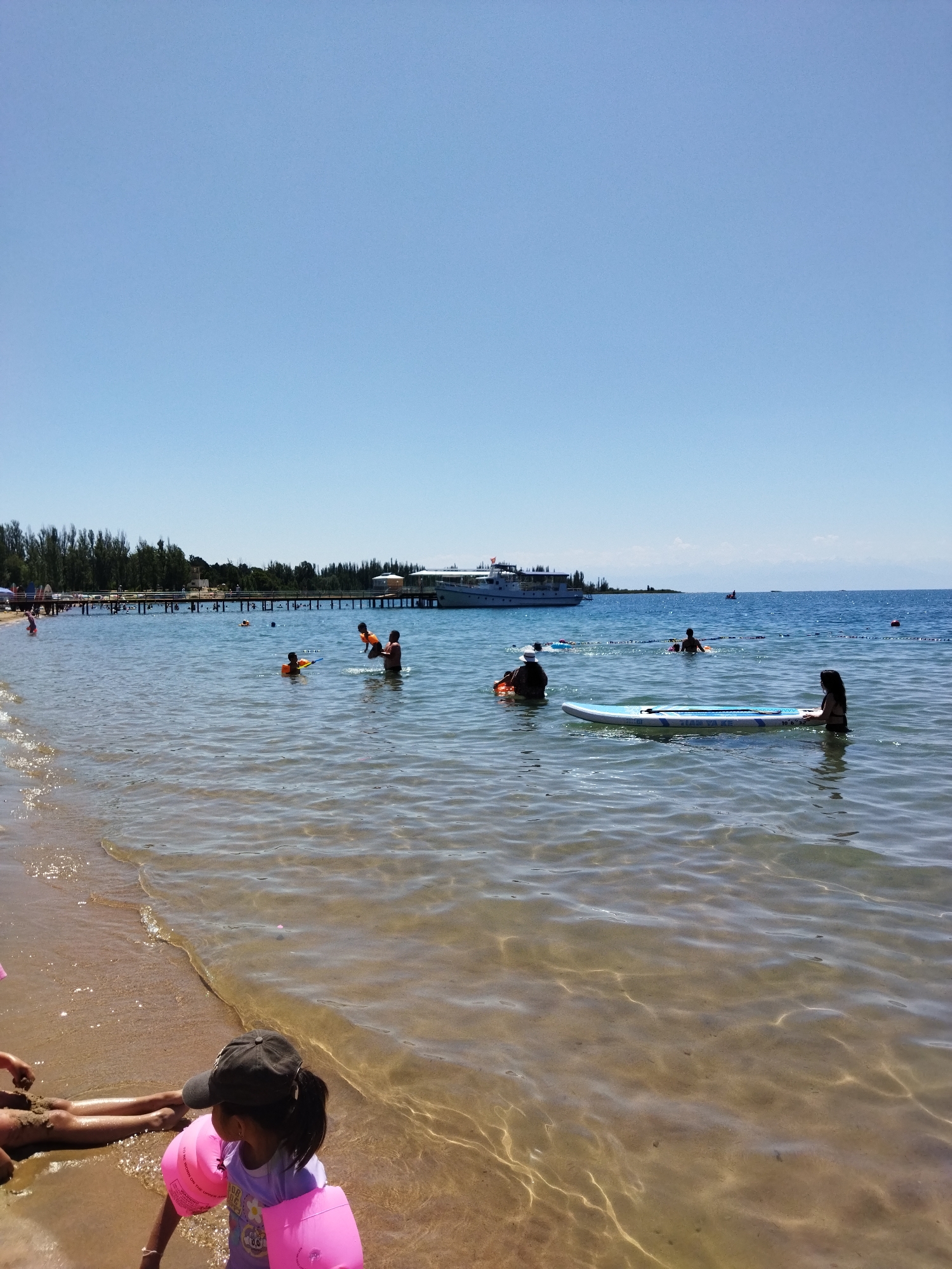 An eye-level, medium shot captures a sunny day at a beach in Kara-Oy, Kyrgyzstan. The calm, clear blue water gently laps at the sandy shore, where a young girl with a dark baseball cap and pink arm floaties is visible. Further out, several people are enjoying the water. One adult is lifting a child, while others swim or float. A paddleboard is visible in the water, with a person sitting on it. In the background, a long wooden pier extends into the water, with a large white boat docked at its end. Trees line the distant shore, and the sky is a clear, bright blue. The overall scene depicts a leisurely summer day at a lakeside beach.