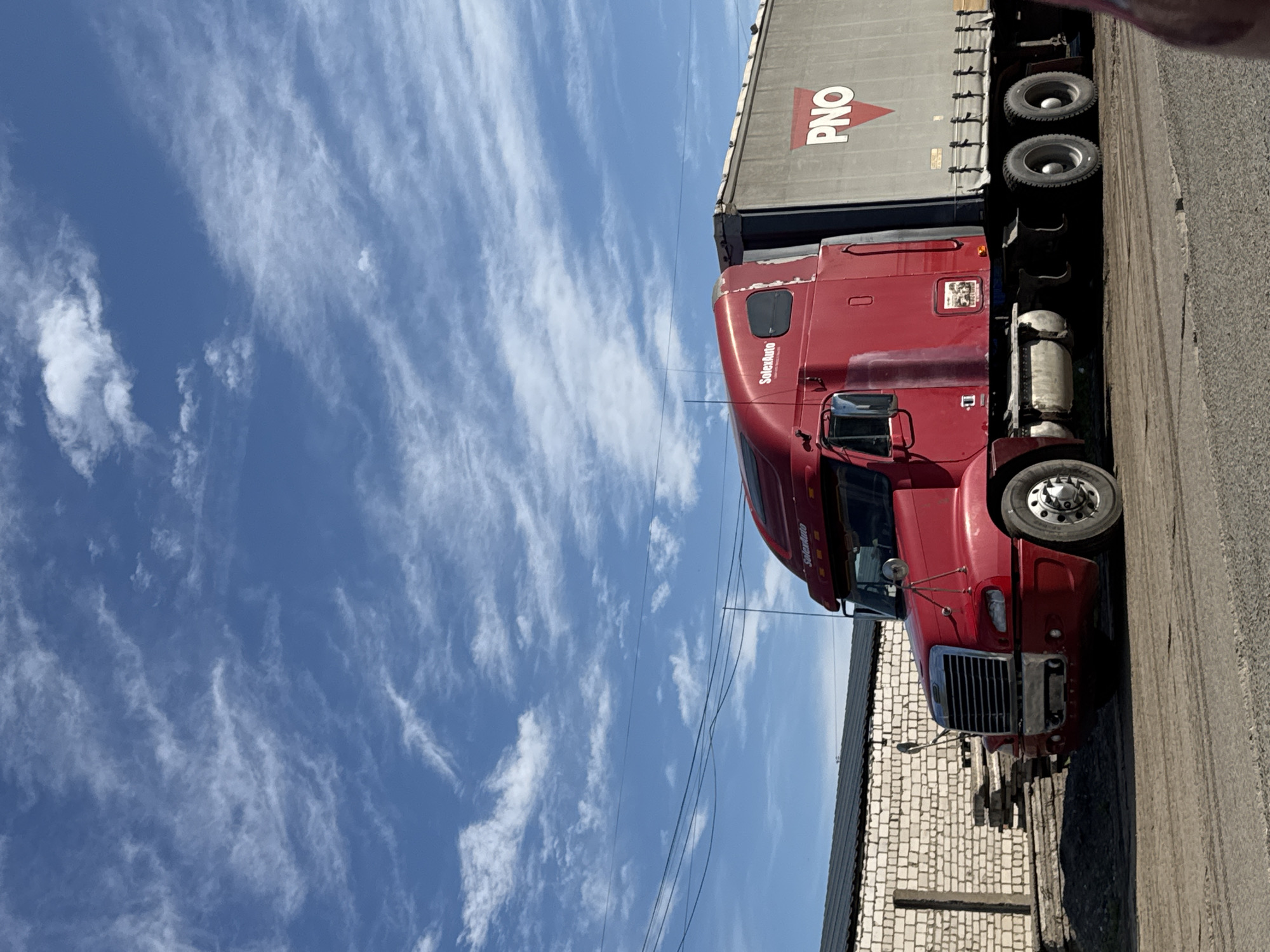 A red Freightliner semi-truck is depicted parked outdoors, likely in an industrial or commercial area in Chaikovsky, Russia. The tractor unit, a deep red color with chrome details, including the front grille and wheel hubs, is connected to a gray semi-trailer. The trailer features a covered top, possibly a tarp system, and prominently displays the "PNO" logo in red with an upward-pointing triangular symbol. On the side of the truck's cab, above the driver's door, the text "SolexAuto" is visible.
The scene is set under a clear blue sky, which fills a significant portion of the image. Scattered wispy white cirrus clouds are visible across the sky, indicating fair weather conditions. The bright and even lighting suggests it is daytime. In the background, a structure with white brick walls can be seen, partially obscured by the truck. Overhead, thin power lines stretch across the frame. The ground beneath the truck appears to be asphalt or a similar paved surface. There are no people visible, and the truck appears stationary, suggesting it is either awaiting activity or at rest.