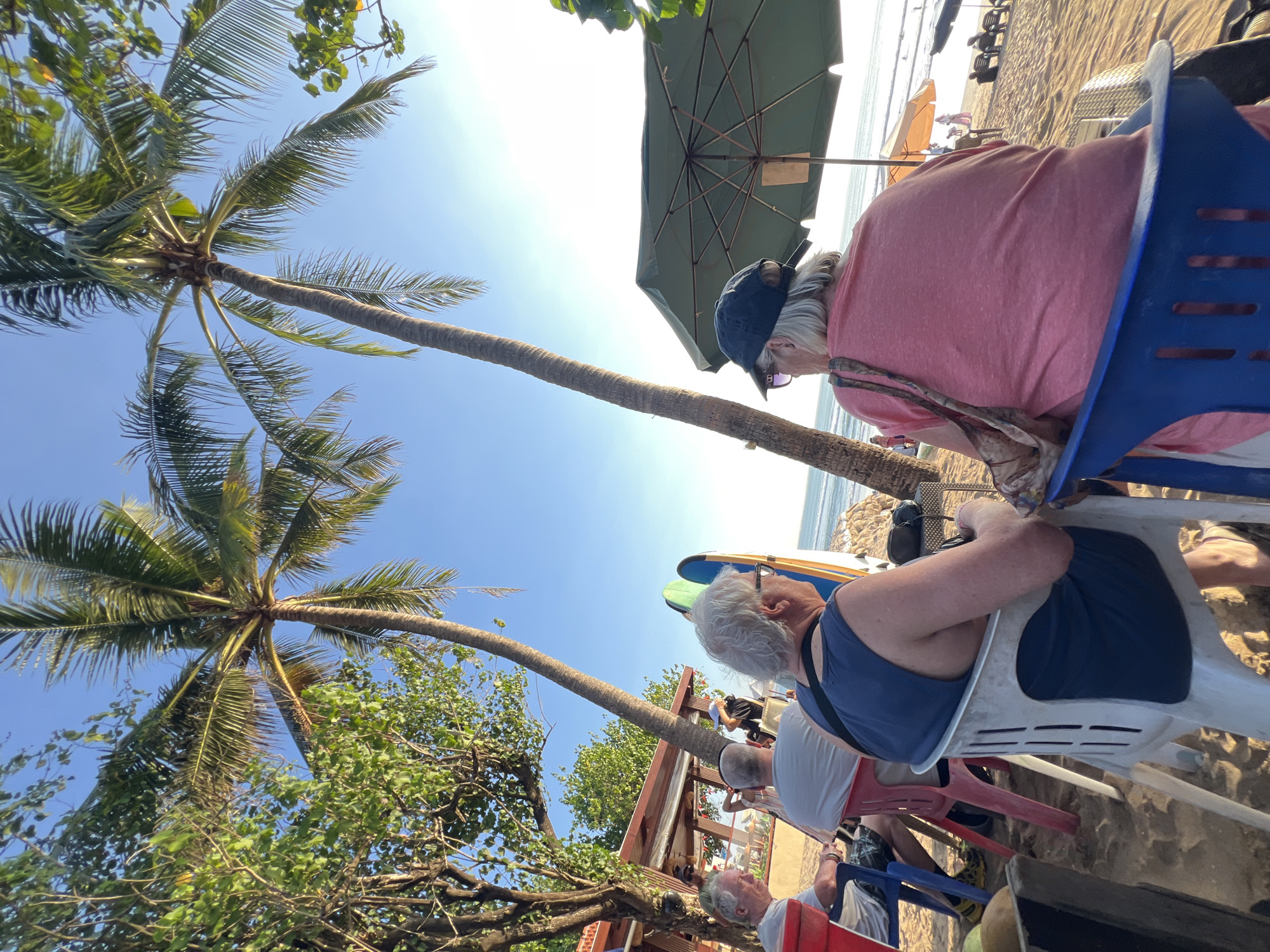 The image depicts a beachfront scene in Kuta, Indonesia, under a clear blue sky. Two mature individuals are seated in chairs facing the ocean. The person in the foreground, viewed from behind, wears a pink t-shirt and a dark baseball cap. To their left, another individual with gray hair and glasses sits in a blue chair, wearing a dark blue tank top. In the background, several other people are visible, some seated and others standing, near beach umbrellas and what appear to be surfboards. Palm trees are prominent in the foreground and middle ground, with lush green foliage framing the scene. The sandy beach stretches towards the ocean, where gentle waves can be seen. A structure resembling a beachside cafe or restaurant is partially visible on the left side of the image. The overall atmosphere suggests a relaxed day at the beach.