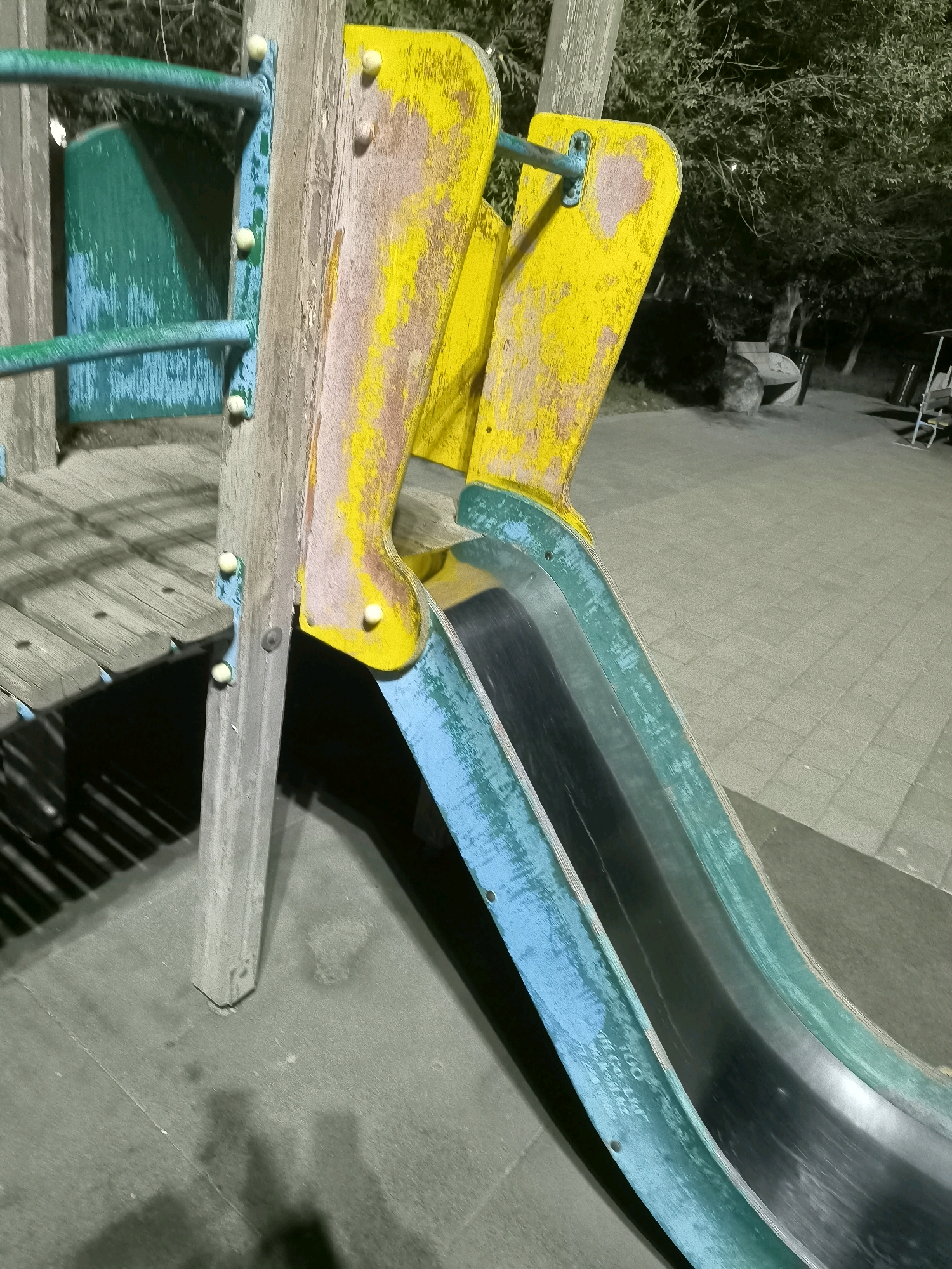 The image shows a close-up view of a playground slide in Jezqazğan, Kazakhstan. The slide is made of metal and plastic, with a green and blue exterior and a black interior. The yellow portion of the slide appears to be wood and is weathered and peeling. The slide is attached to a wooden support structure. In the background, there are trees and a paved area. The lighting suggests it is either evening or nighttime, with artificial lights illuminating the scene.