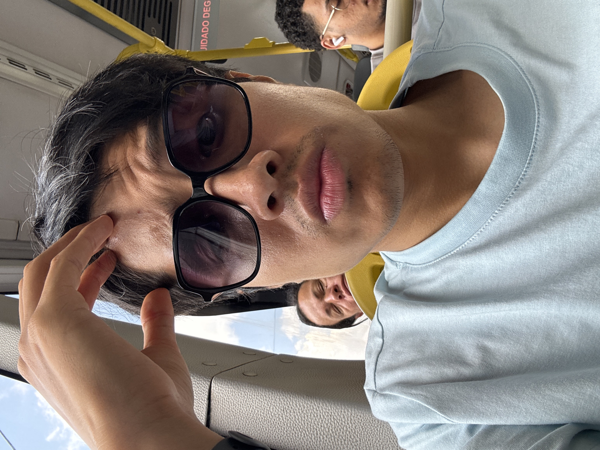 A man wearing dark sunglasses and a light blue t-shirt is looking directly at the camera, with his hand touching his forehead. The scene is inside a public transportation vehicle, likely a bus, based on the yellow handrails and the presence of other passengers in the background. The vehicle is in the city of São Paulo, Brazil, evidenced by the signage with the word "CUIDADO" (Portuguese for "Caution").