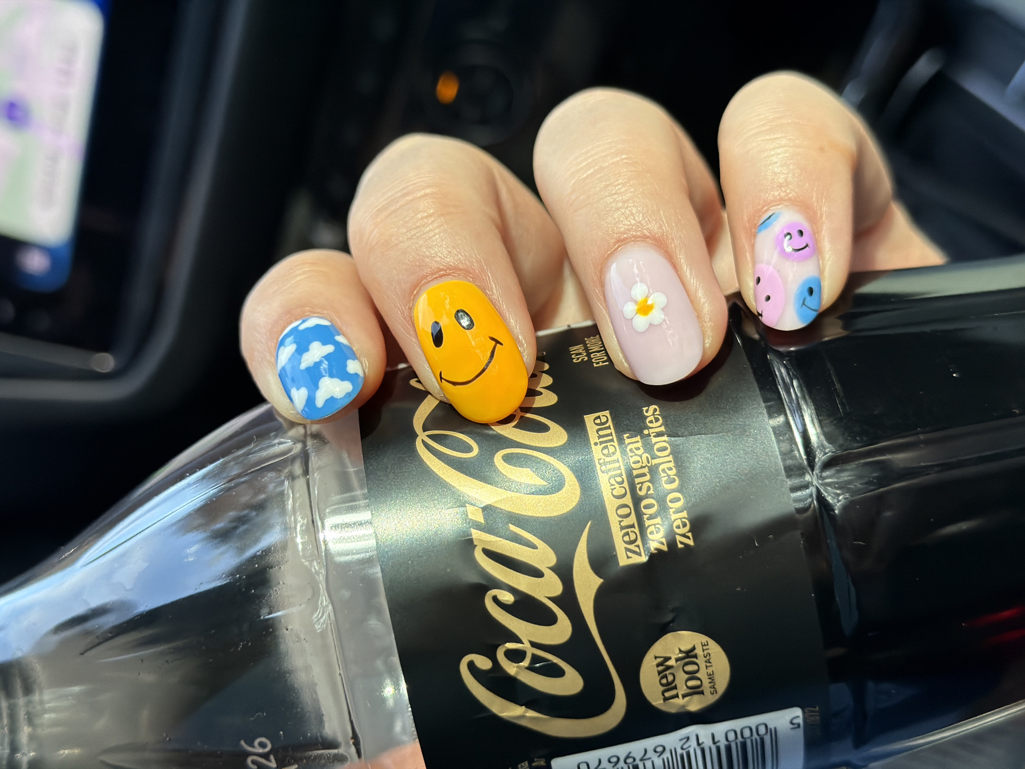 A close-up shot shows a hand holding a plastic bottle of Coca-Cola Zero. The focus is on the hand and the bottle, revealing several details. The hand's fingernails are painted in various playful designs: one features blue with white clouds, another a yellow smiley face, a pale pink nail with a white flower, and a pink nail adorned with small pink and blue smiley faces.
The Coca-Cola bottle is dark with gold lettering, indicating "Coca-Cola zero caffeine zero sugar zero calories." A label on the bottle states "new look SAME TASTE." A barcode is also visible at the bottom of the label.
The background is dark and indistinct, except for a small area showing a blurred screen, likely of a navigation device, suggesting the scene is set inside a vehicle.
Based on the OCR result there is no city of Berģi, Latvia visible in the image.