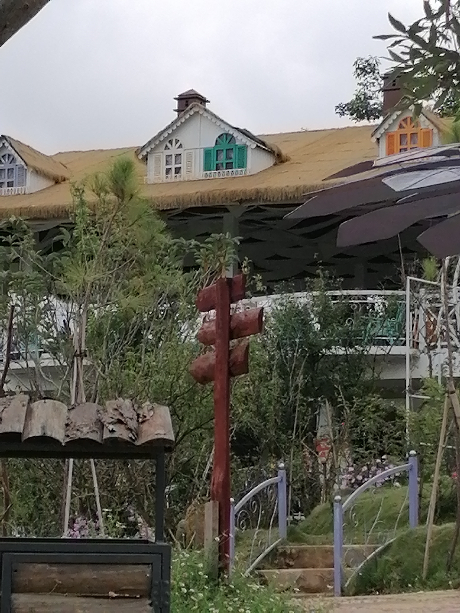 This image captures a picturesque building nestled within lush greenery under an overcast sky in Lạc Dương, Vietnam. The multi-story structure features white walls and a distinctive, possibly thatched or straw-covered, light brown roof. Several dormer windows punctuate the roofline, each with white frames and brightly colored shutters: light blue on the far left, vibrant green in the center, and orange on the right. A small chimney is visible above the rightmost dormer.
Below the roof, a white-railed balcony suggests outdoor seating or viewing areas, partially obscured by dense green foliage. In the immediate foreground, a rustic, reddish-brown wooden signpost stands centrally, featuring three roughly shaped, blank wooden signs pointing in different directions. To the lower left, a decorative wooden structure, possibly a well cover or stand, with curved log elements and a dark metal base is visible. On the right, a small, arched pedestrian bridge with light blue/lavender metal railings and stone steps winds up a grassy incline. Various bushes and small trees, including a slender pine-like tree, contribute to the verdant garden setting. A large, dark, multi-panel awning or umbrella partially covers the right side of the building.
The scene conveys a tranquil, whimsical atmosphere, characteristic of a resort, guesthouse, or themed cafe. No people are visible, and there is no discernible text in the image. The overcast weather suggests daytime.