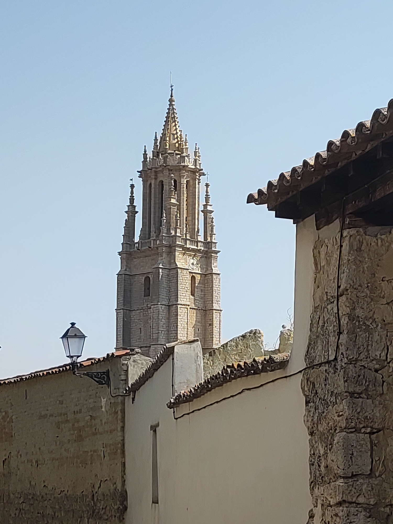 The image shows a view of the bell tower of the Collegiate Church of Santa María la Real in Ampudia, Spain, from a street level perspective. The bell tower is a tall, Gothic-style structure made of light-colored stone, with intricate carvings, arched windows, and multiple spires. It stands against a clear blue sky. In the foreground, there are rooftops and stone walls of buildings, with a tiled roof and a traditional wrought-iron lantern on the left. The stone walls are textured and weathered, suggesting an old architectural style. The overall scene depicts a historic urban landscape bathed in daylight. There are no people visible in the image.