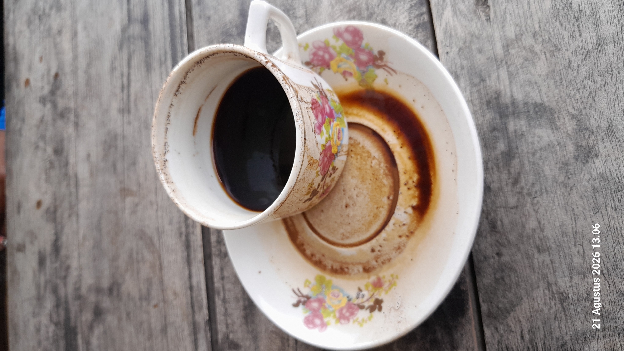 A close-up, overhead shot shows a white ceramic coffee cup with a floral pattern sitting on a matching saucer. The cup contains dark coffee, and the saucer has a ring of coffee residue and grounds from where the cup was previously placed. The items are situated on a weathered, gray wooden surface, possibly a table. The timestamp "21 Agustus 2026 13.06" is visible in the bottom left corner. The scene is likely set in an outdoor or rustic indoor environment in Takeran, Indonesia, given the timestamp. There are no people visible in the image.