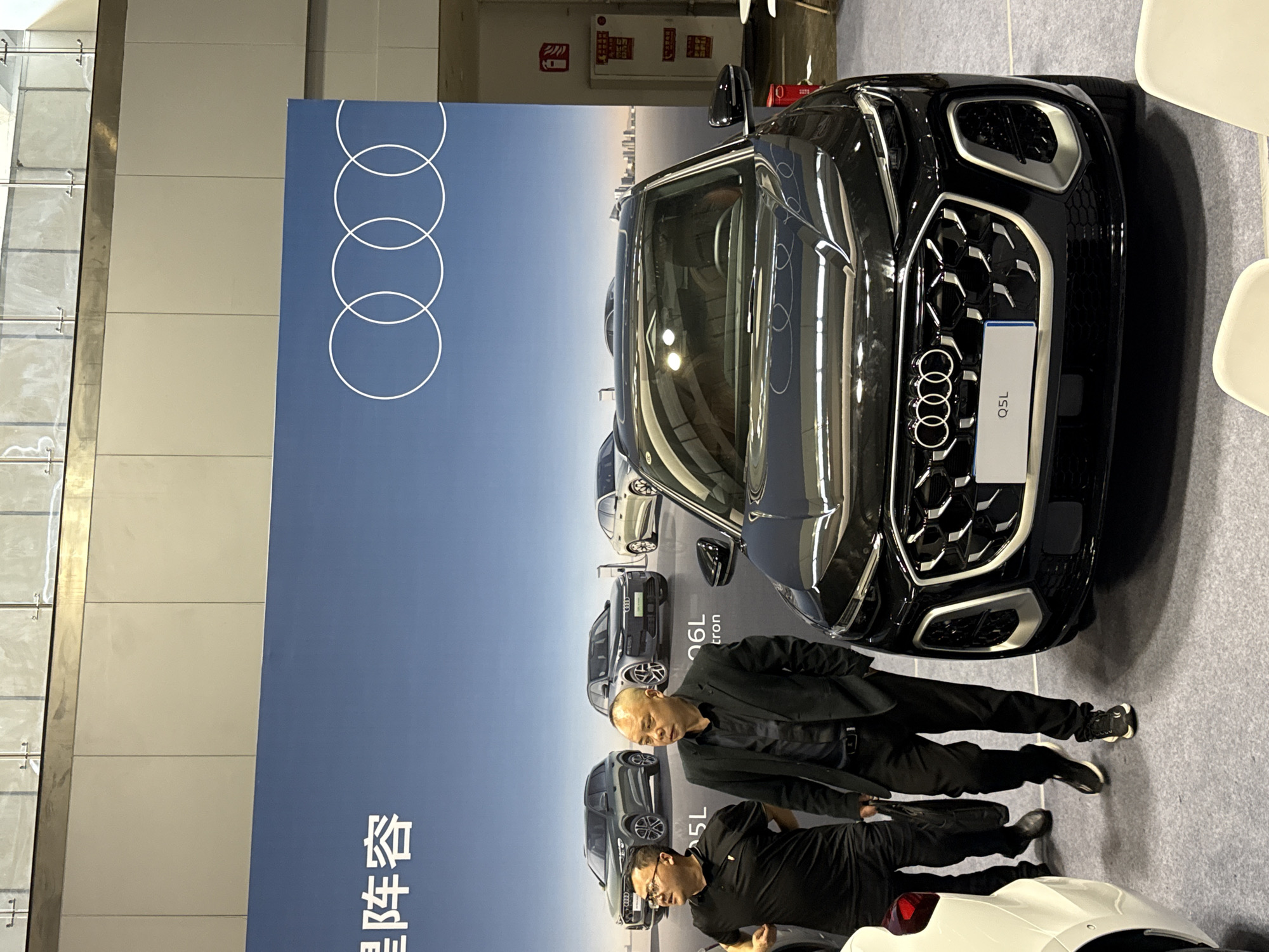 The image shows a black Audi Q5L in front of a promotional banner with the Audi logo and the car model printed on it. Two men are standing next to the car. The background includes parts of an interior space with windows and walls. The location is not identifiable.