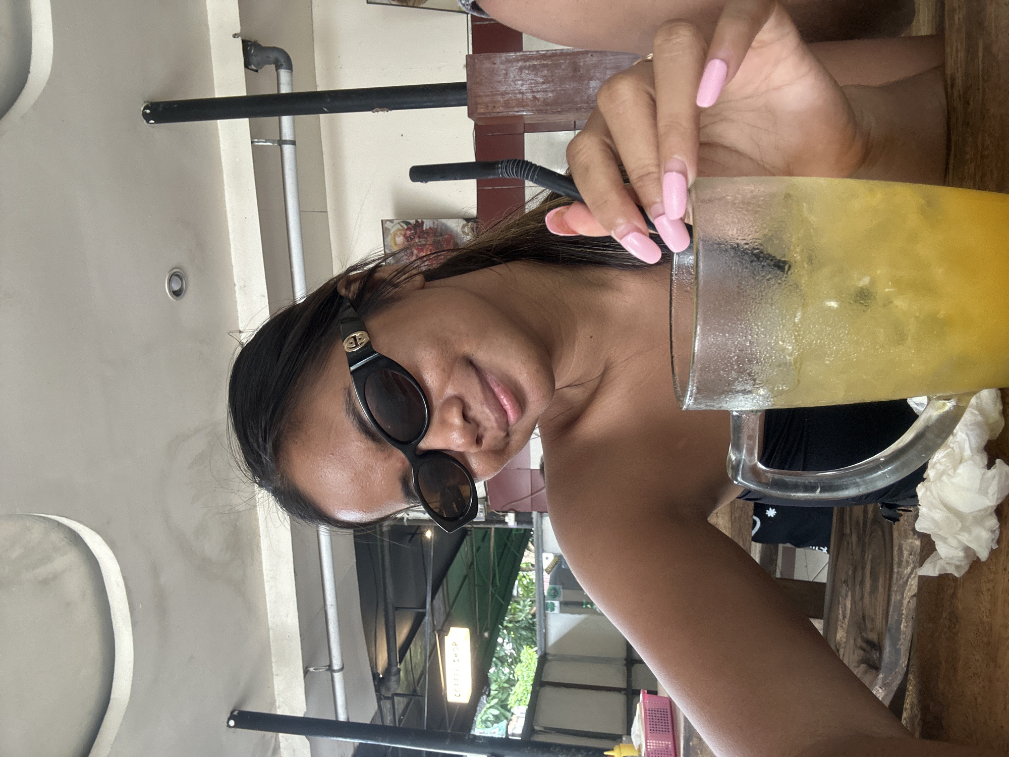 A young woman is taking a selfie at a table in a cafe in Kuta, Indonesia. She is wearing black sunglasses, and a black strapless top. She has long dark hair and light-colored skin. Her nails are painted a light pink color. She is holding a straw in a glass of yellow liquid with ice. The table is made of wood. In the background, there are signs, and greenery. The ceiling is made of concrete.