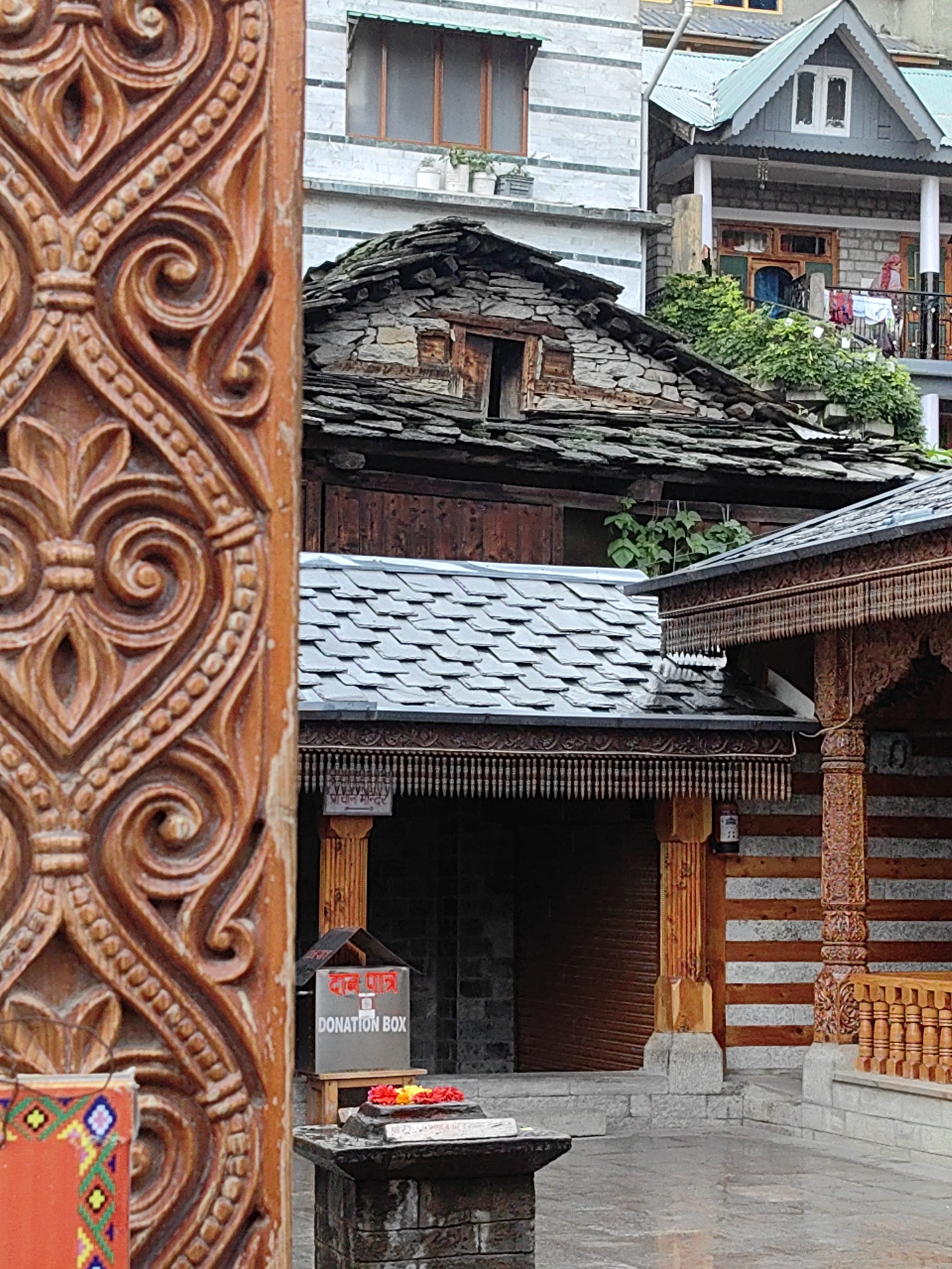 The image depicts a street scene in Vashisht, India, focusing on the intricate wooden carvings of a doorway in the foreground. Beyond the doorway, traditional Himachali architecture is visible, characterized by stone and wood structures with slate roofs. A donation box is placed on a stone pedestal, adorned with red and yellow flowers. The environment appears to be a courtyard or a pathway, with stone paving. In the background, multiple buildings with varied architectural styles and roofing materials are stacked along a hillside. The overall impression is one of a culturally rich and architecturally distinct locale.