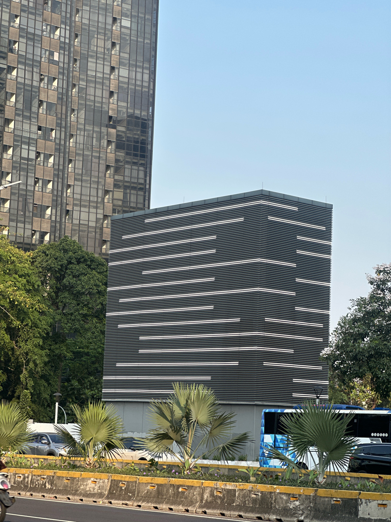 The image depicts a street scene in South Jakarta, Indonesia, with a focus on modern architecture and urban infrastructure. In the background, a tall, glass-covered skyscraper with a grid-like facade dominates the left side of the frame, reflecting the sky and surrounding environment. To its right stands a more contemporary, dark-colored building with a distinctive facade characterized by numerous horizontal white lines, creating a visual pattern.
In the foreground, a road is visible with a concrete barrier separating it from a median strip. The median strip is adorned with palm trees and low-lying vegetation, suggesting a tropical urban setting. Vehicles are present on the road, including a silver sedan and a bus with a blue and white geometric pattern. The sky is clear and blue, indicating fair weather. The overall impression is one of a bustling city with a mix of old and new architectural styles, set against a backdrop of lush greenery.