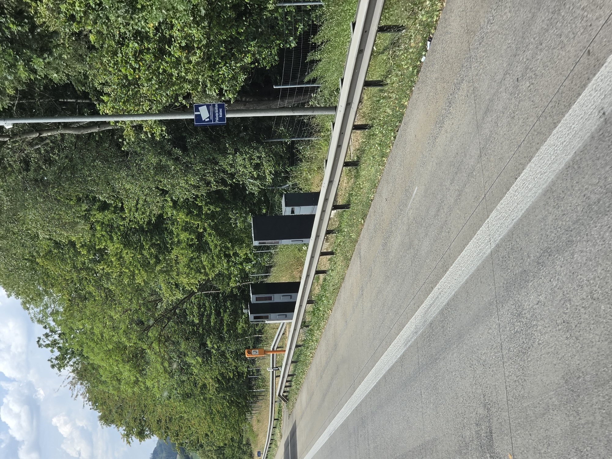 The image shows a road with multiple speed cameras on the side, situated near a densely wooded area. A guardrail separates the road from the cameras and the vegetation. A sign with a camera icon and text "Polizeipräsidium Koblenz" is visible on a pole, indicating that this is a monitored area by the Koblenz police. The road appears to be a highway or a major road, with clear lane markings. The trees are lush and green, suggesting it is summertime. The sky is partly cloudy. There are no people visible in the image. The overall scene suggests a location on a road where speed enforcement is in place.