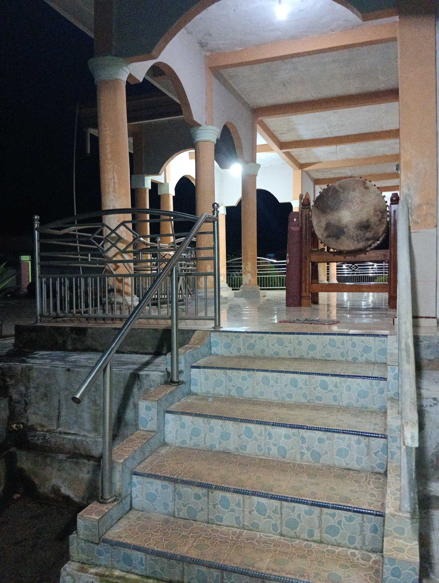 The image depicts an outdoor stairway leading up to a covered area with pillars, likely a part of a mosque or religious building in Pematang Jaya, Indonesia. The stairway has light blue tiled risers and stone treads, with a metal handrail on the right side. The covered area has large, rounded, light brown pillars and an open ceiling with exposed beams. In the background, a large drum, possibly a bedug used for calling to prayer, is visible, resting on a wooden stand. The overall scene is dimly lit, suggesting it is nighttime or dusk, with some artificial lighting present within the covered area. There are no people visible in the image.