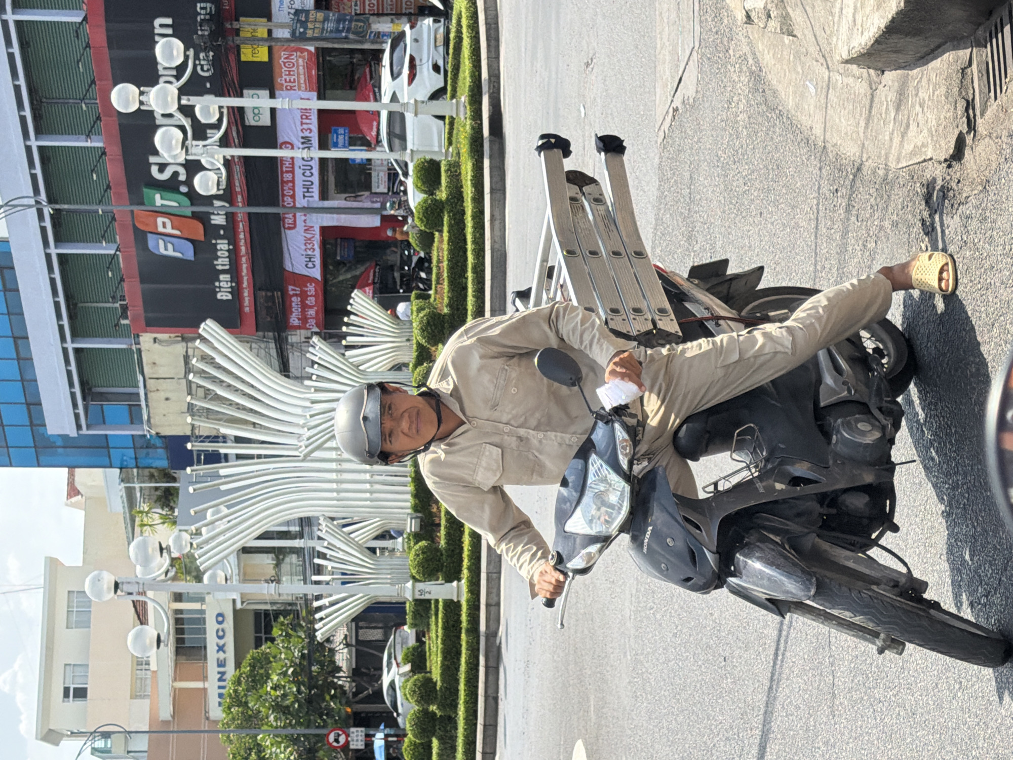 A man wearing a light tan uniform and a dark helmet sits on a black Honda scooter in an outdoor urban environment, likely a street intersection, in Nha Trang, Vietnam. He is transporting a folded, multi-section ladder secured to the rear of the scooter, extending to the right of the frame. The scooter has a black body and a silver kickstand. The man's left hand is on the handlebar, and his right hand is near the ladder. He wears light yellow sandals.
In the background, a large white sculpture with multiple vertical, curved elements is visible, as are trees, buildings with signage, and other vehicles. One building features a prominent sign for "FPT Shop". Traffic lights and street lamps are also present. The scene appears to be during daylight hours, with a bright sky and distinct shadows.