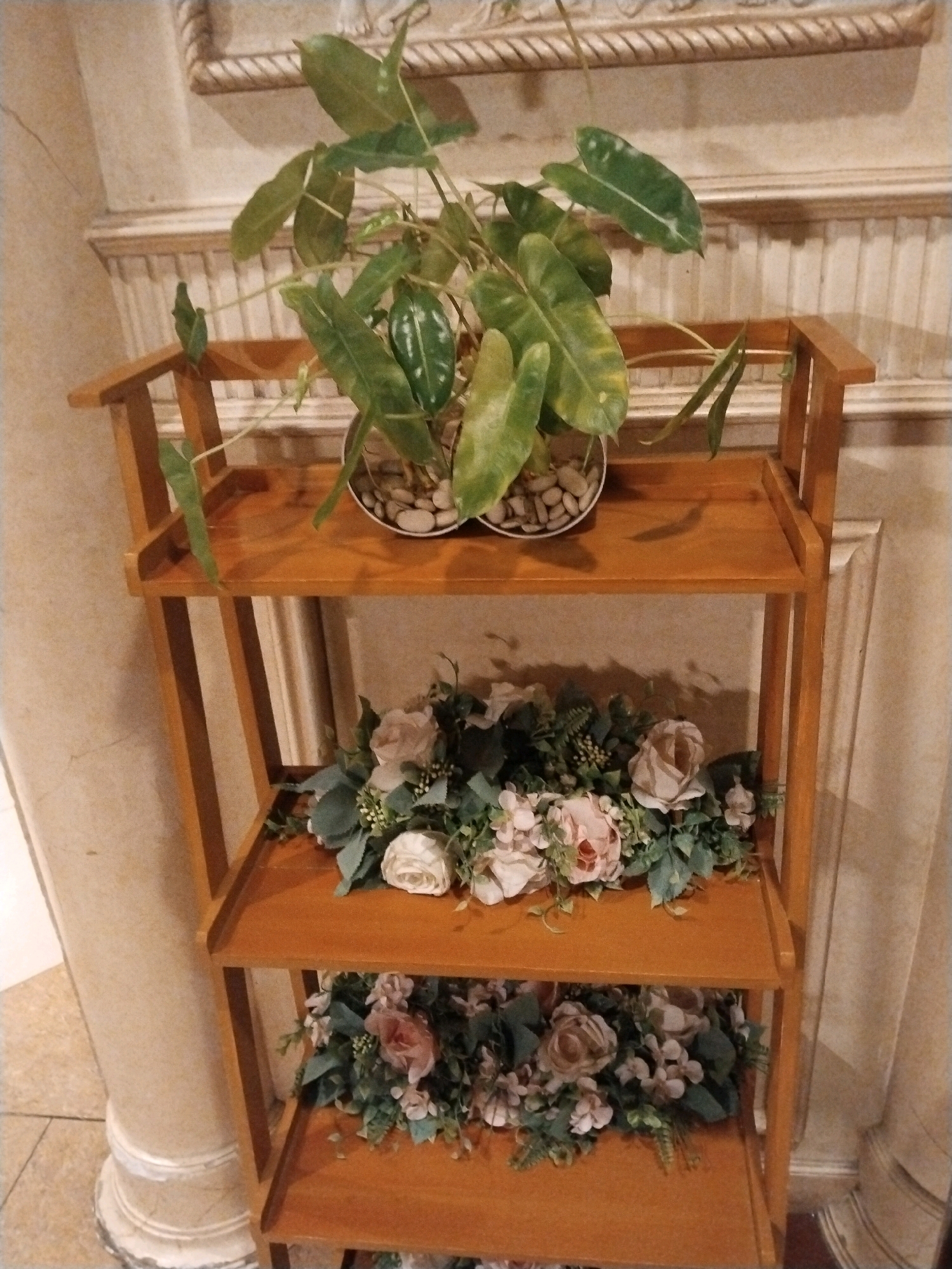 A wooden three-tiered shelf unit is positioned against a wall with decorative molding and vertical slats. On the top shelf sits a potted green plant with large, elongated leaves and pebbles in the pot. The middle shelf and bottom shelf are adorned with artificial floral arrangements consisting of roses and other greenery in muted pinks and creams. The scene appears to be indoors, possibly in a home or event space, given the decorative elements of the wall and the presence of the shelf unit. The location in Sinduadi, Indonesia, is not discernible from the image.
