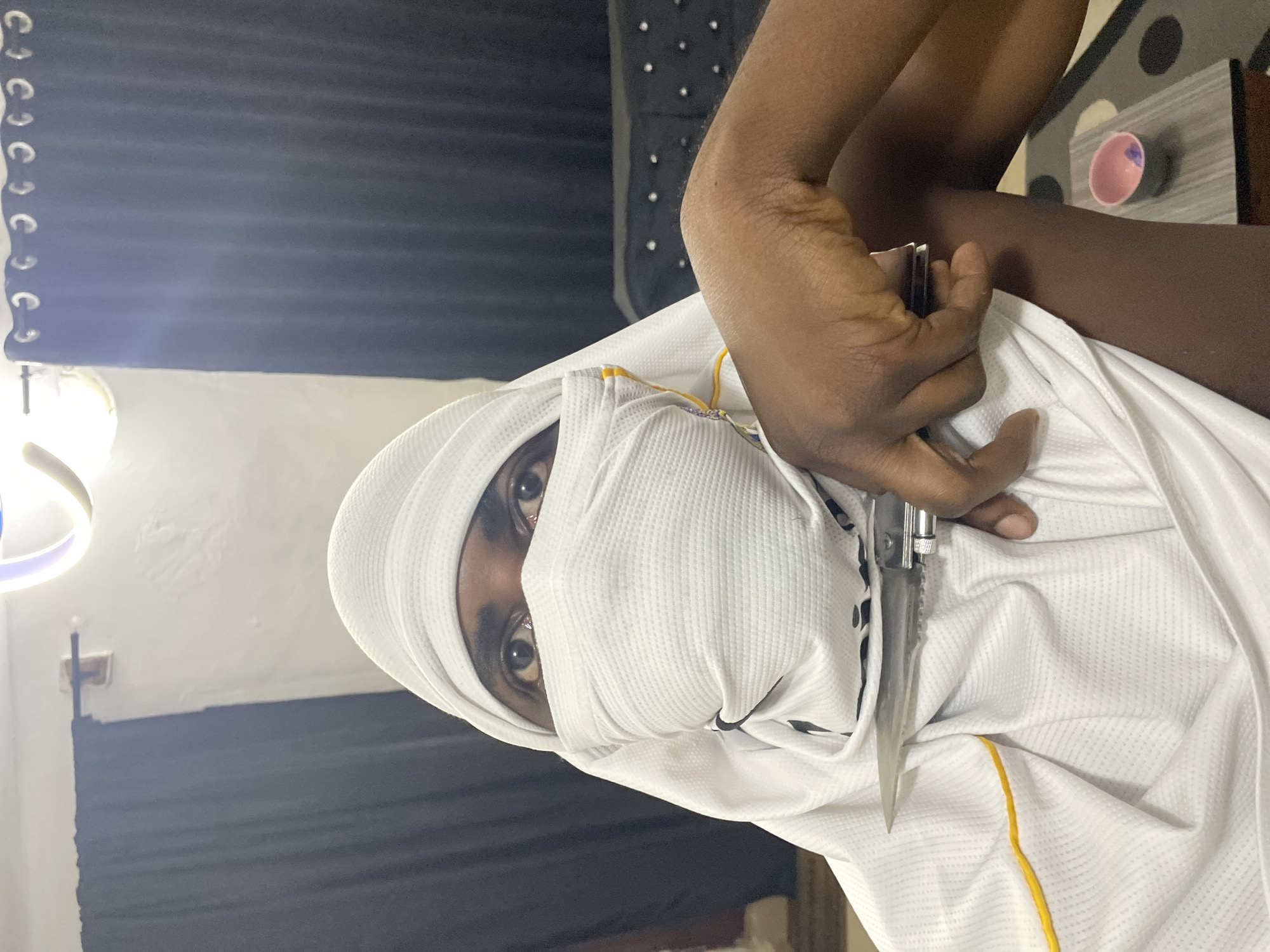 A person wearing a white head covering and mask is holding a knife to their arm. The person is indoors, with dark curtains in the background. There is a light fixture on the ceiling and a table with a small bowl on it. The person's eyes are visible, and they appear to be looking at the camera. The image does not contain information that could place the location in Akwanga, Nigeria.