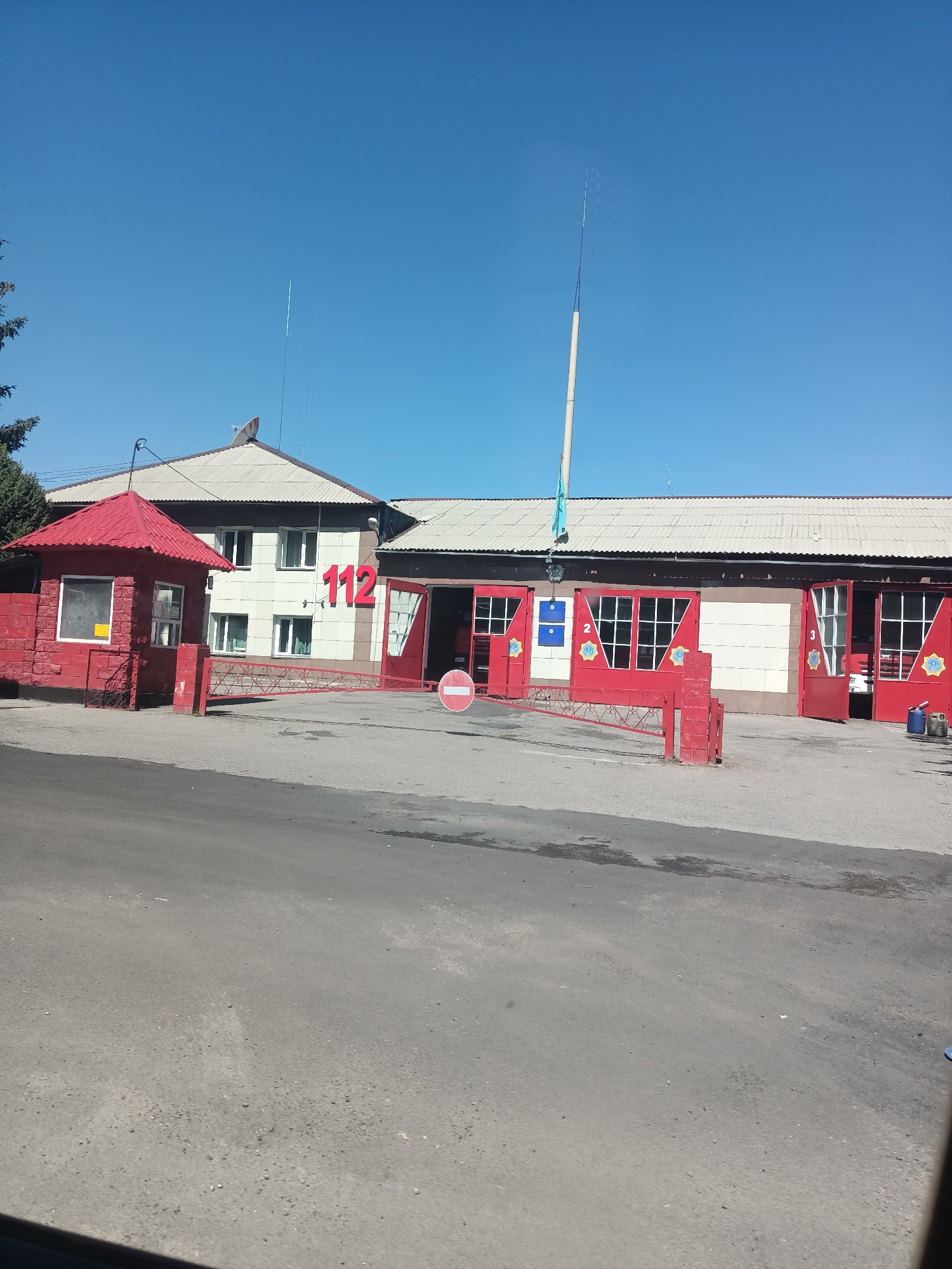 The image depicts the exterior of a fire station located in the city of Talgar, Kazakhstan, under a clear blue sky. In the foreground, a paved road leads towards the building. To the left, a small red guardhouse with a glass window is visible, and in front of it, a red brick wall. A red gate separates the road from the fire station complex.
The fire station itself is a long, light-colored building with a corrugated roof. Large red garage doors are a prominent feature, with three doors clearly visible. Two of these doors are open, revealing glimpses of the interior. The number "112" in large red numerals is displayed on the front of the building. The numbers "2" and "3" are also visible on the garage doors. A flagpole with a flag is present on the roof, along with some antennas. A red sign with a white circle and a diagonal line, indicating "no entry," is placed on the right side of the gate. Various objects, including a blue bucket and a red container, are scattered near the open garage doors. A satellite dish is visible on the roof of the main building. The overall scene suggests a functional fire station in a dry, sunny environment.
