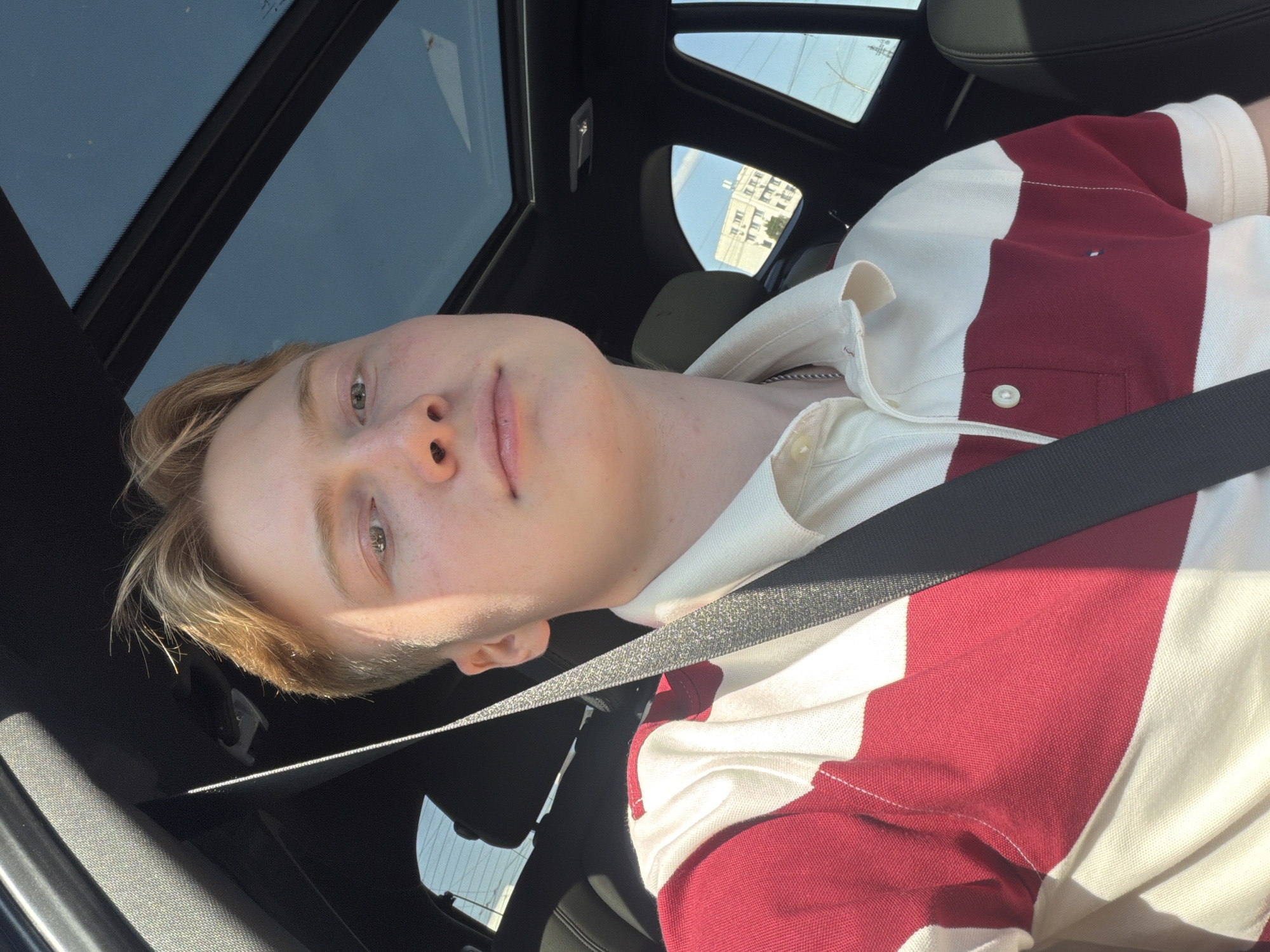 A young man with blond hair is captured in a selfie from the chest up, seated in the driver's seat of a car. He is wearing a red and white striped collared shirt. Sunlight streams through the car's open sunroof, illuminating his face and casting shadows on his clothing. The background reveals the interior of the car, including a seatbelt across his chest and a portion of the car's dashboard and roof. Through the windows, a blue sky with some clouds and buildings can be seen, suggesting an urban environment. The presence of buildings and the sky implies the location is likely outdoors in a city, however, the specific location in Kyiv, Ukraine cannot be determined from the visual information alone.