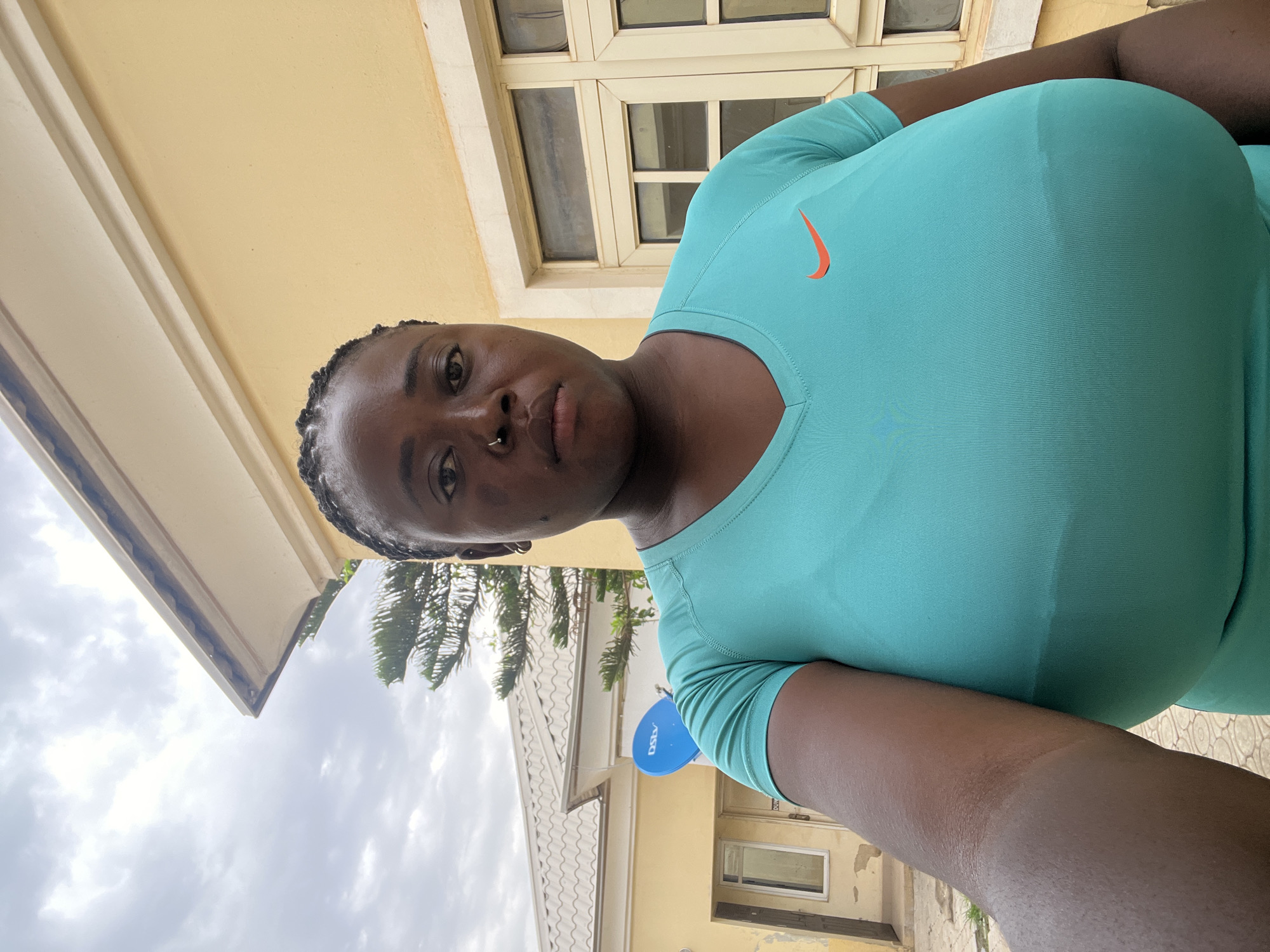 A close-up, medium shot shows a Black woman with dark skin and braided hair from the chest up, facing the camera. She is wearing a teal Nike t-shirt with an orange Nike logo on the right side of her chest. She has a nose ring and a small hoop earring in her left ear. Her expression is neutral. Behind her, a light yellow building with white trim and windows is visible, along with a satellite dish on the roof of a smaller, white-roofed building. The sky is overcast and appears cloudy. The image was taken outdoors, likely in Abuja, Nigeria, based on the presence of a DStv satellite dish, which is common in Nigeria. The scene depicts a casual, everyday moment.