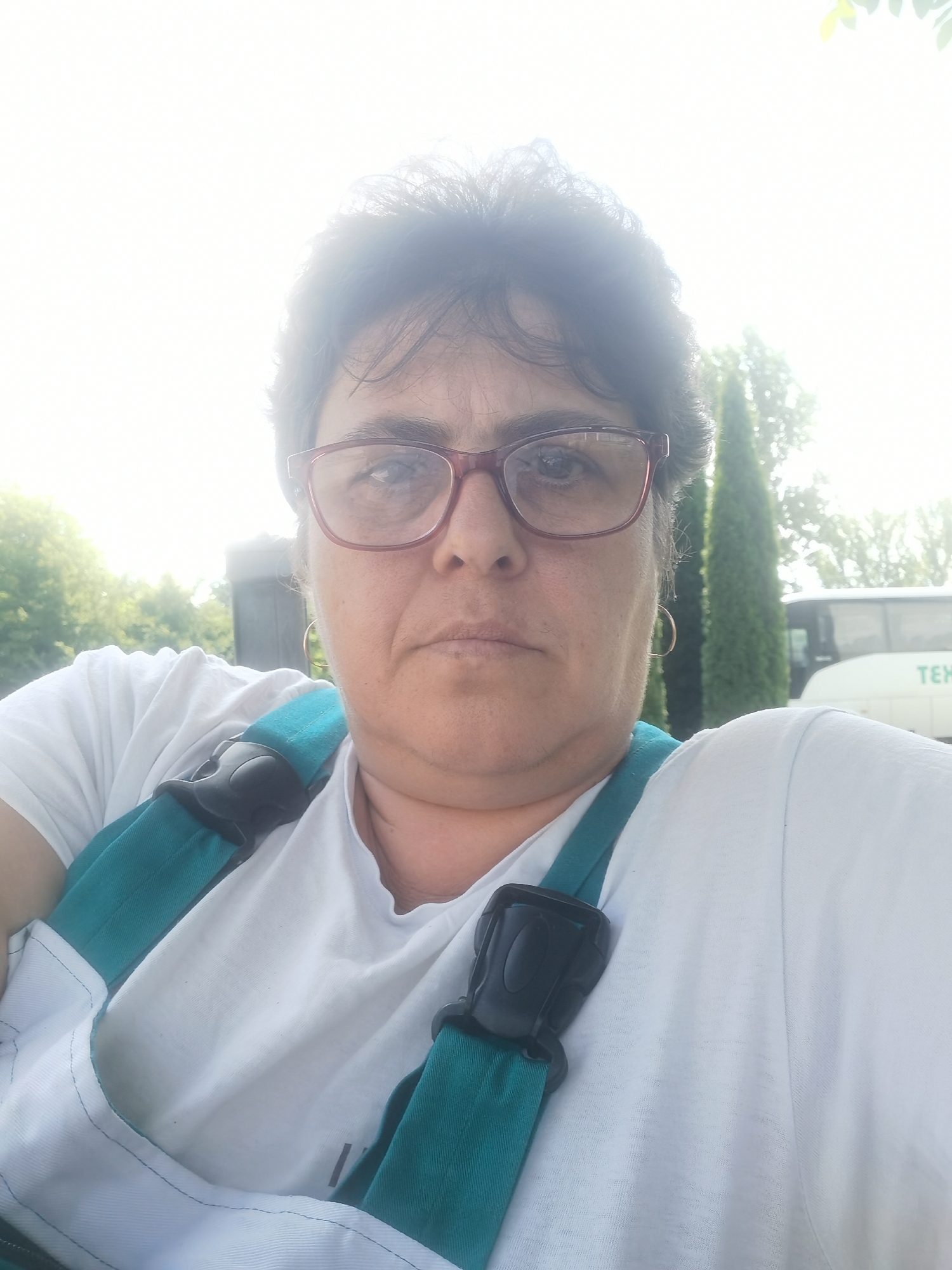 A selfie taken outdoors shows a woman with dark hair and glasses. She is wearing a white t-shirt and a teal-colored vest with black buckles. Behind her, there are trees and a white bus with "TEX" visible on its side. The lighting suggests it is daytime, possibly with bright sunlight. The background appears to be a park or an area with greenery and a road. There is no indication of the specific location within Lomets, Bulgaria.