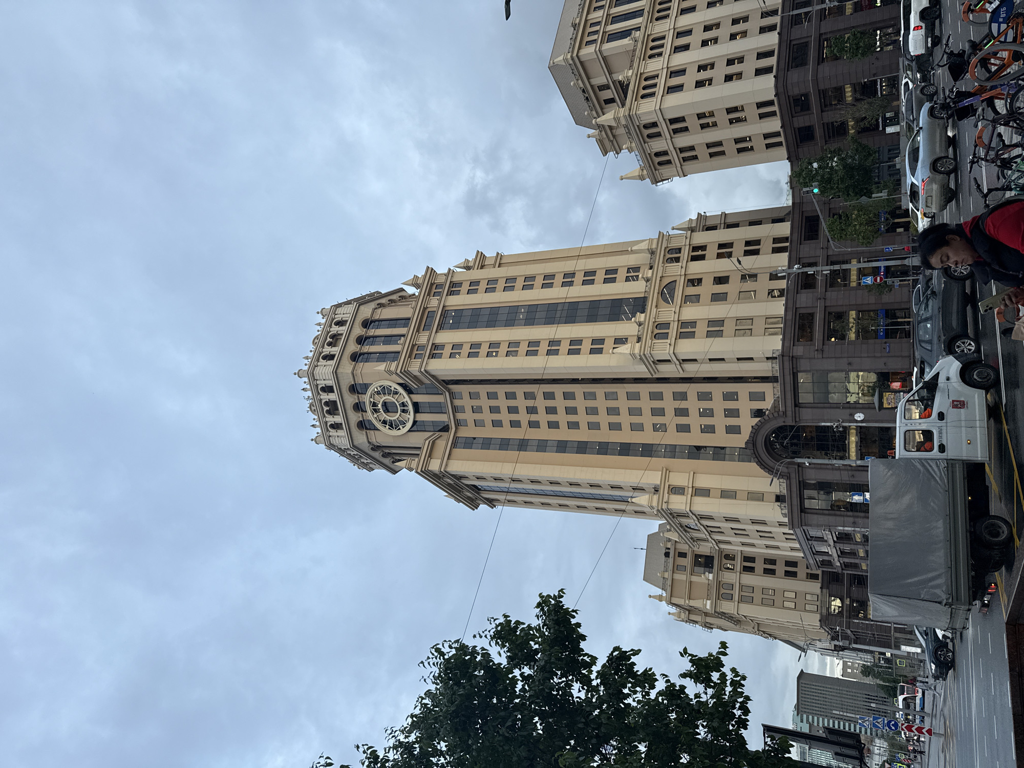 The image captures a tall, ornate building with a prominent clock face, situated in Moscow, Russia. The building is multi-storied, with a light-colored facade and numerous windows arranged in a grid pattern. The architectural style suggests a modern interpretation of classical designs.
The street scene in front of the building includes parked cars, bicycles, and a delivery truck. A person is visible near the sidewalk, holding a mobile phone. Green trees partially obscure the view of the street-level buildings. The sky is overcast, indicating a cloudy day.