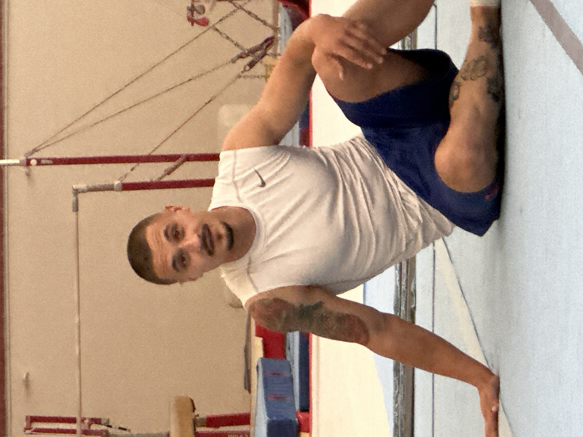 A man is performing a gymnastic stretch in a gymnastics facility. He is wearing a white t-shirt with a Nike logo on the left chest, and blue shorts. He has a tattoo on his left arm and another on his right leg. His right leg is bent and against a white wall. His right arm is extended to brace himself against the wall. Gymnastics equipment, including uneven bars, is visible in the background. The location is consistent with a gymnastics training facility. The specific location within Sofia, Bulgaria cannot be determined from this image.