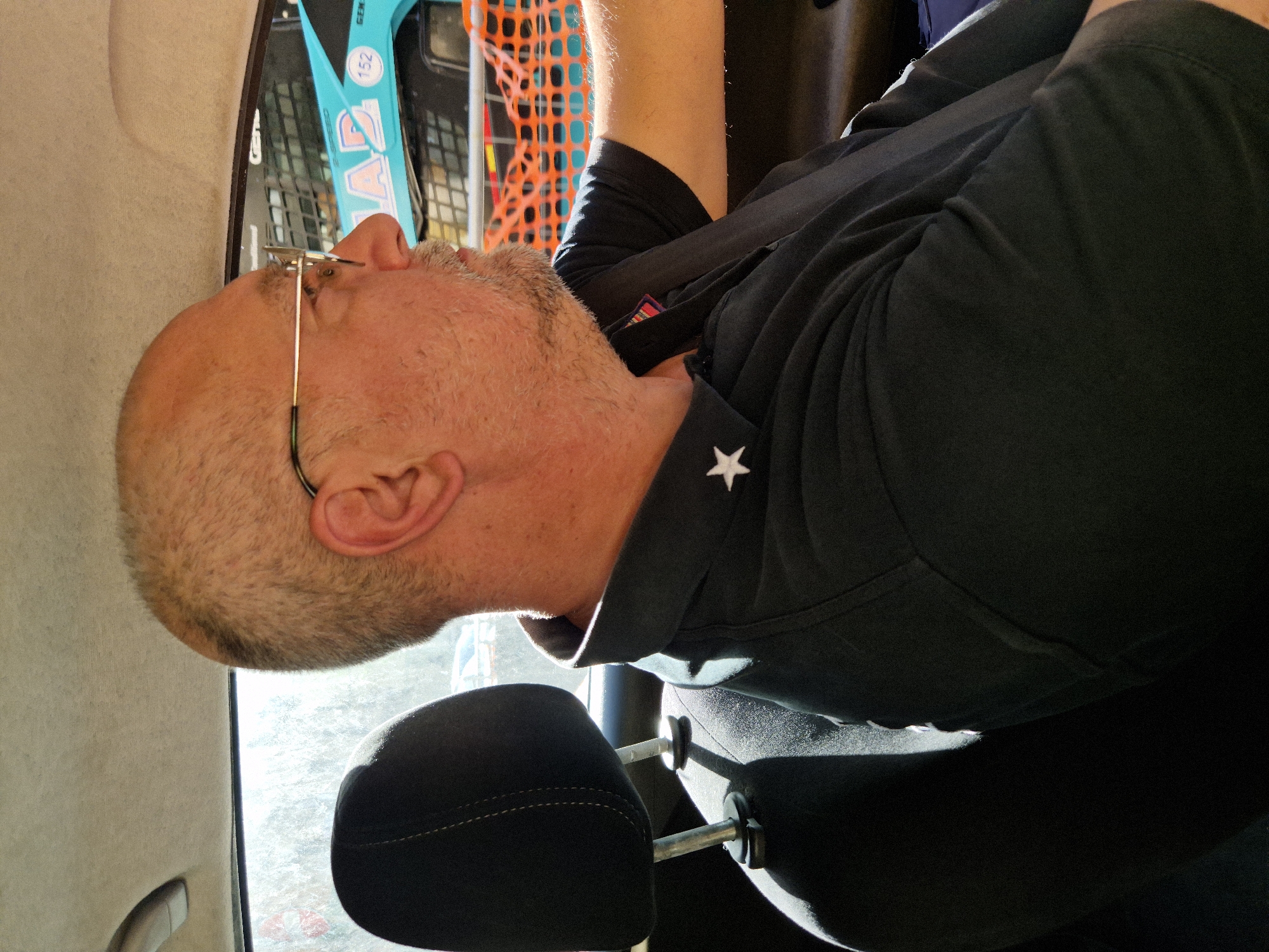 A middle-aged Caucasian man with a shaved head, wearing glasses and a dark polo shirt with a white star emblem, is seen in profile from the chest up, sitting in the passenger seat of a vehicle. Sunlight illuminates his face and neck from the side. Outside the vehicle, a construction site with orange netting and a blue vehicle with the text "TAD 152" is visible. The car interior features a black headrest and a beige headliner. The scene is likely taking place within the industrial or port area of Taranto, Italy, given the construction and signage.