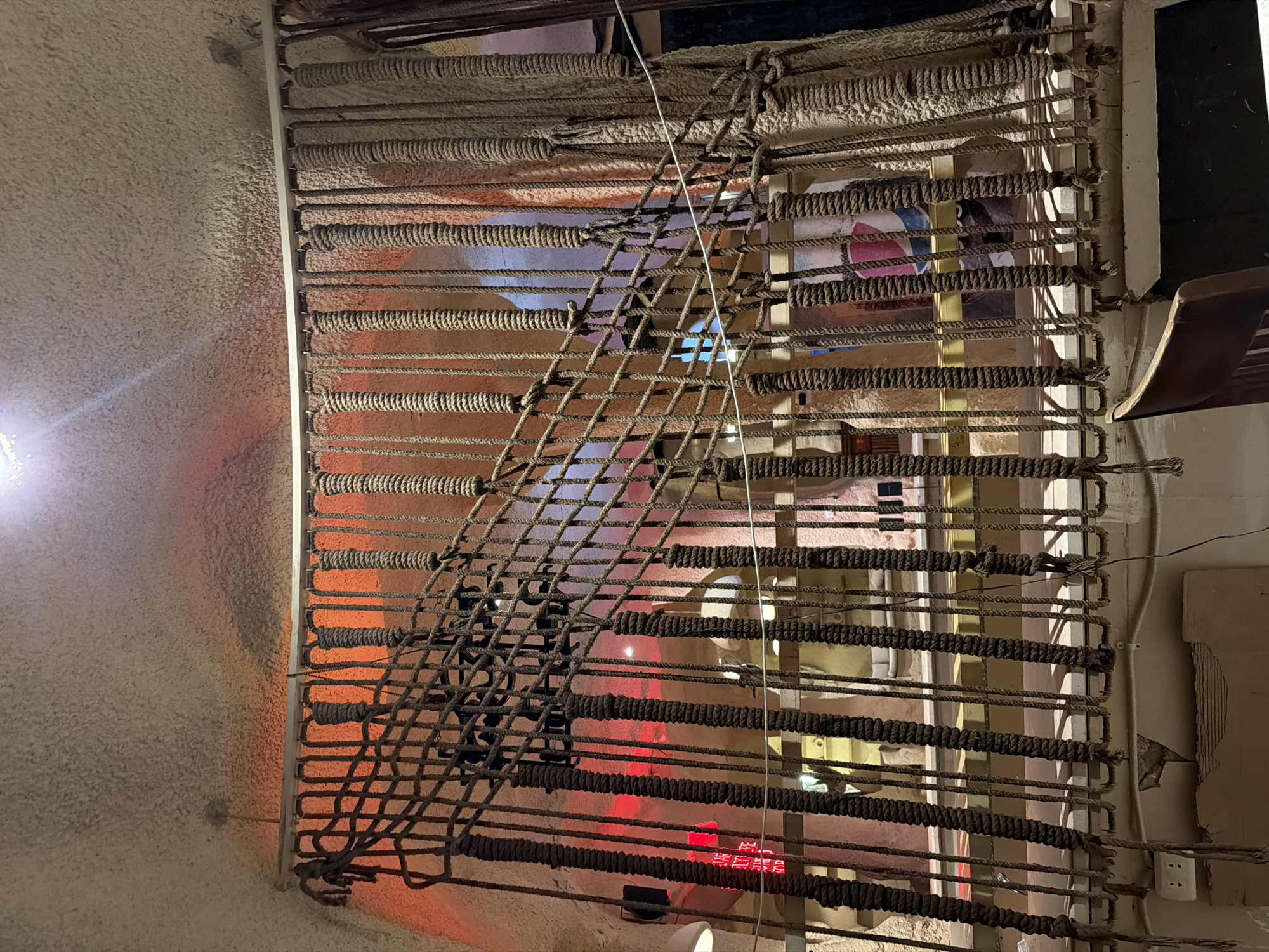 The image depicts the interior of a dimly lit establishment, possibly a restaurant or bar, with a rustic and somewhat industrial aesthetic. The dominant feature is a railing made of thick, tightly wound ropes, creating a vertical barrier. A net woven from similar rope is draped across this railing, adding to the textured and somewhat haphazard design. Behind the rope railing, a staircase is visible, also seemingly constructed with wooden steps and metal supports, leading upwards. The background reveals an uneven, possibly stucco or concrete wall with an orange-red ambient light source illuminating it, casting shadows and creating a warm, albeit dramatic, atmosphere. Scattered within the background are various objects, including what appear to be tables and chairs, hints of signage with red neon lettering, and decorative elements that contribute to the overall ambiance. A ceiling light fixture is visible at the top of the frame, casting a focused beam downwards. The overall impression is of a unique and potentially atmospheric indoor space, possibly designed to evoke a specific theme. The location is identified as Sharm El Sheikh, Egypt.