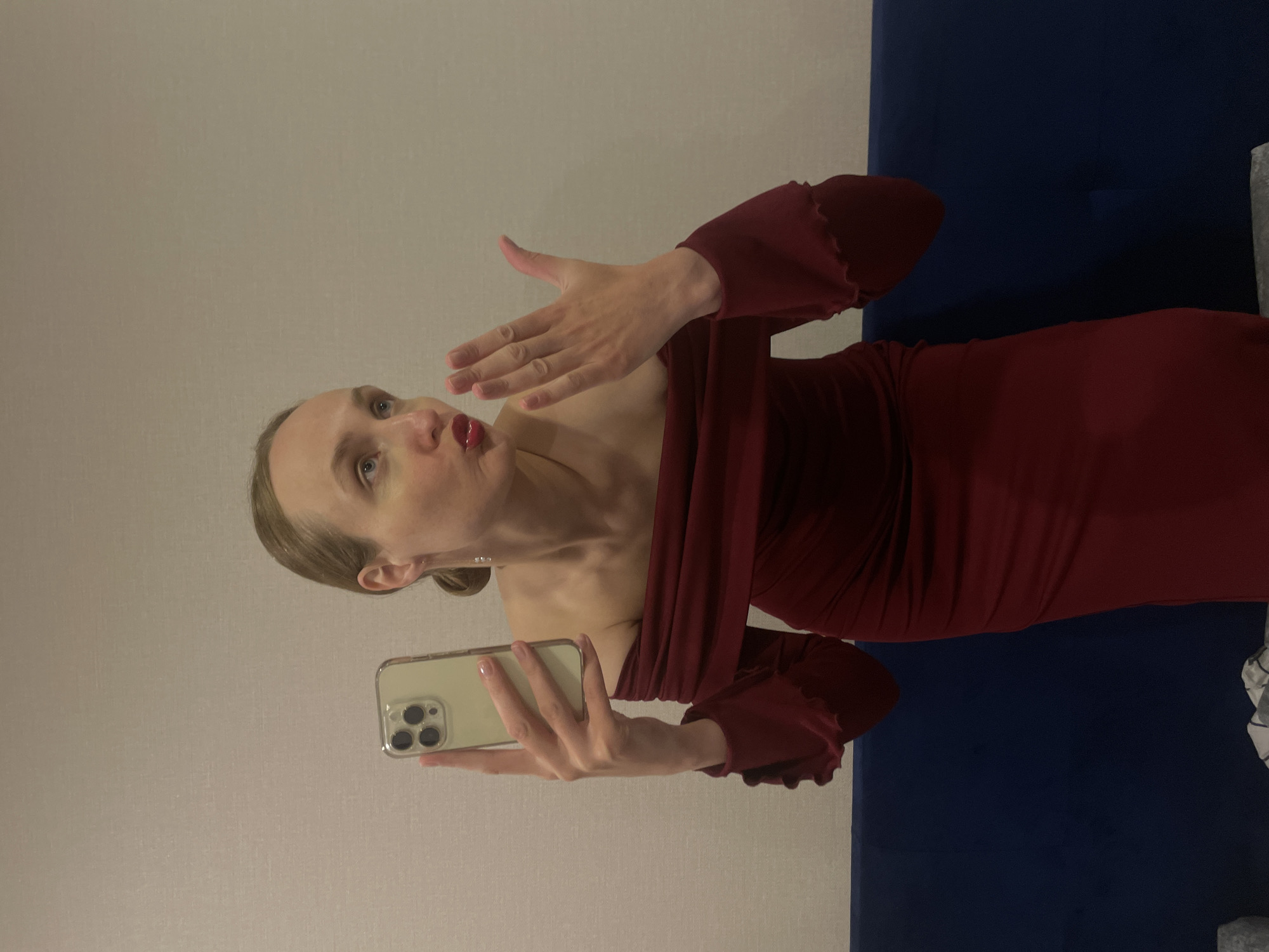 A woman is taking a selfie in what appears to be an indoor setting, possibly a hotel room, given the plain wall and a dark blue headboard or furniture piece behind her. She is wearing a red, off-the-shoulder dress with ruffled sleeves. She is holding an iPhone, likely using it to take the photo, and her left hand is raised, blowing a kiss toward the camera. Her hair is pulled back, and she has makeup on, including red lipstick. The location given is Подшивалово, Russia, but there is nothing in the image to confirm this; it could be any interior setting.