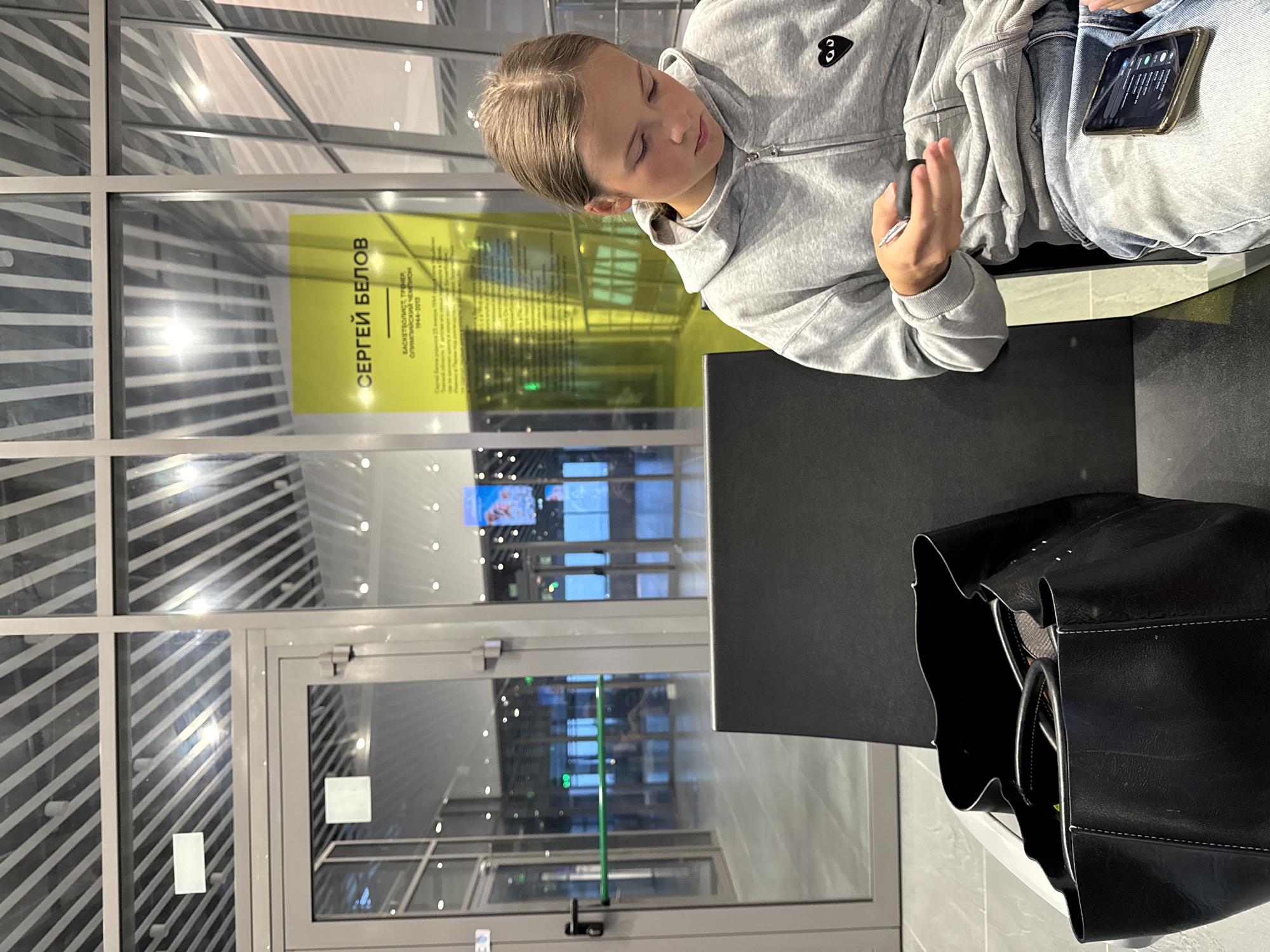 A young Caucasian female with light brown hair, wearing a grey zip-up hoodie with a black heart-shaped logo and blue jeans, is seated and looking down at her smartphone, which is resting on her lap. She holds a small black object in her right hand. Beside her, to the left, is a large black leather tote bag. The background features large glass windows and doors, with the reflection of lights and what appears to be an exhibition display with yellow signage and text, including the Russian words "СЕРГЕЙ БЕЛОВ" and "БАСКЕТБОЛИСТ, ТРЕНЕР, ОЛИМПИЙСКИЙ ЧЕМПИОН 1944-2013", suggesting the location is the Bolshoye Savino International Airport in Russia, potentially in a waiting area or terminal. The lighting is artificial.