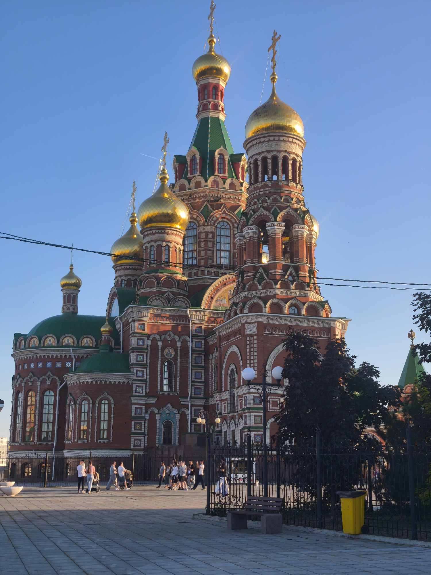 This is a wide, low-angle, exterior shot of the Resurrection Cathedral in Yoshkar-Ola, Russia. The cathedral is an Orthodox church constructed of red brick with green roofs and gold domes. The main section of the cathedral has multiple domes, towers, and intricate architectural details. In the foreground, a paved walkway is visible, with several people walking and pushing strollers. A black metal fence separates the walkway from the cathedral grounds, and trees are visible behind the fence. A park bench and a yellow trash can are also present in the foreground. The sky is clear and blue.