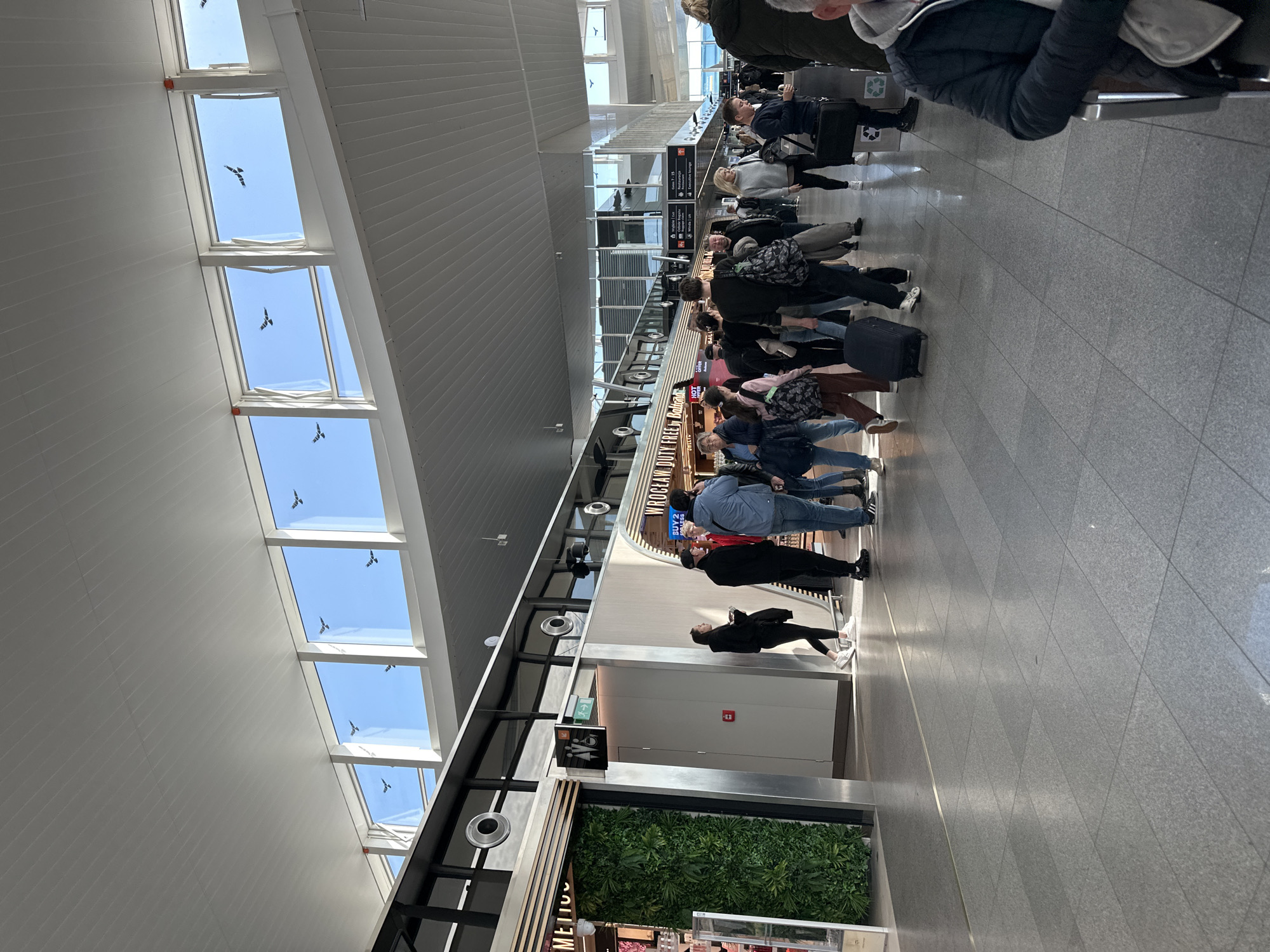 The image depicts a scene inside Wrocław Airport in Wrocław, Poland. People are standing in lines within the terminal. Several individuals have luggage. Overhead, skylights are visible showing a bright blue sky with what appears to be several birds in flight. A shop with green wall decor is visible. There are directional signs present above the line of people. The floor is tiled.