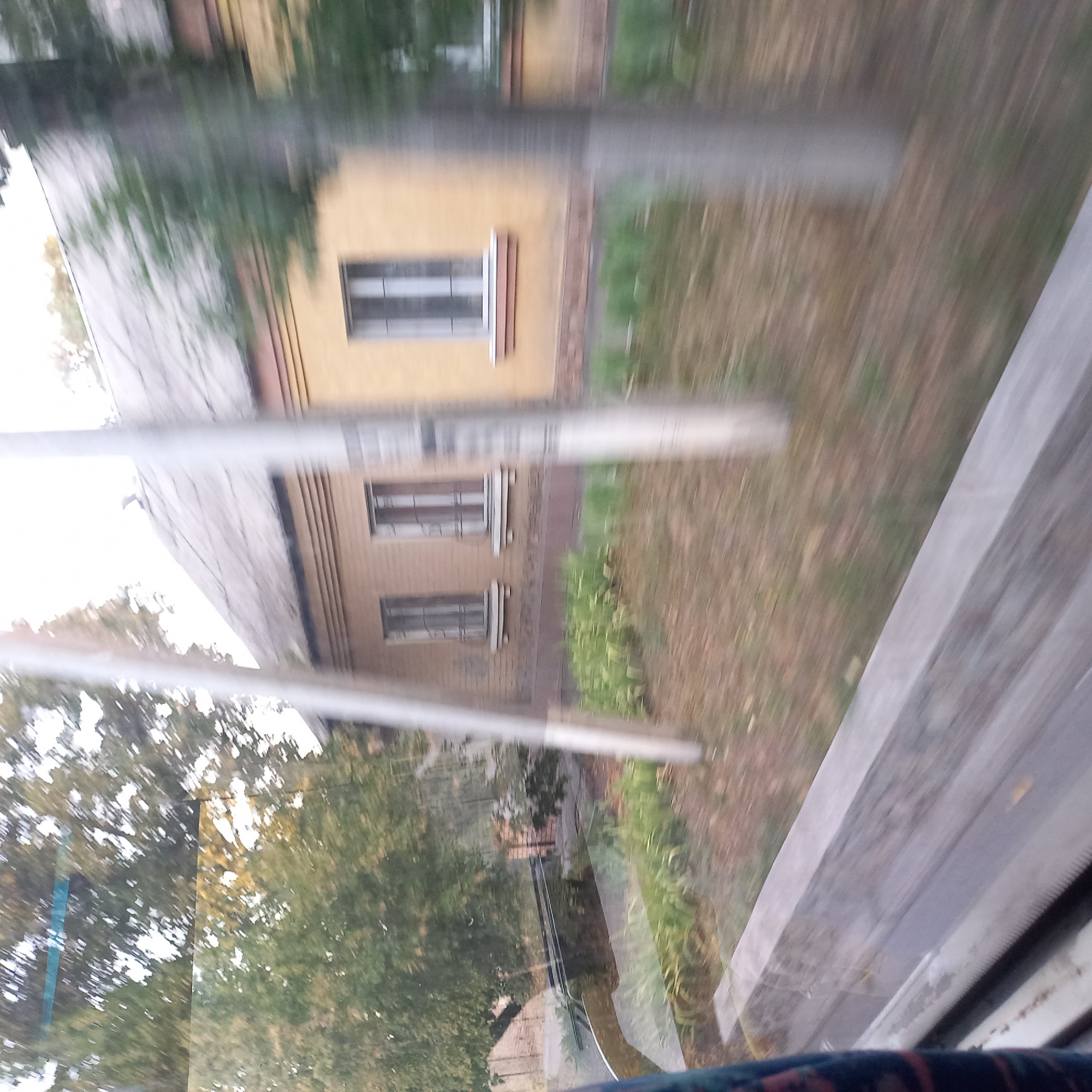 This image, taken from a moving vehicle in Kropyvnytskyi, Ukraine, depicts a two-story building with a light yellow facade and a grey, corrugated roof. The building has multiple windows, some with decorative white sills and metal bars. In front of the building, there is a strip of green vegetation bordering a path or lawn. Trees with green and yellow foliage are visible to the left and right of the building, suggesting an outdoor, possibly suburban or semi-rural, environment. The foreground shows a grey, textured surface that appears to be part of the vehicle from which the photo was taken, possibly a window frame or guardrail. The overall scene is slightly blurred due to the motion of the camera.