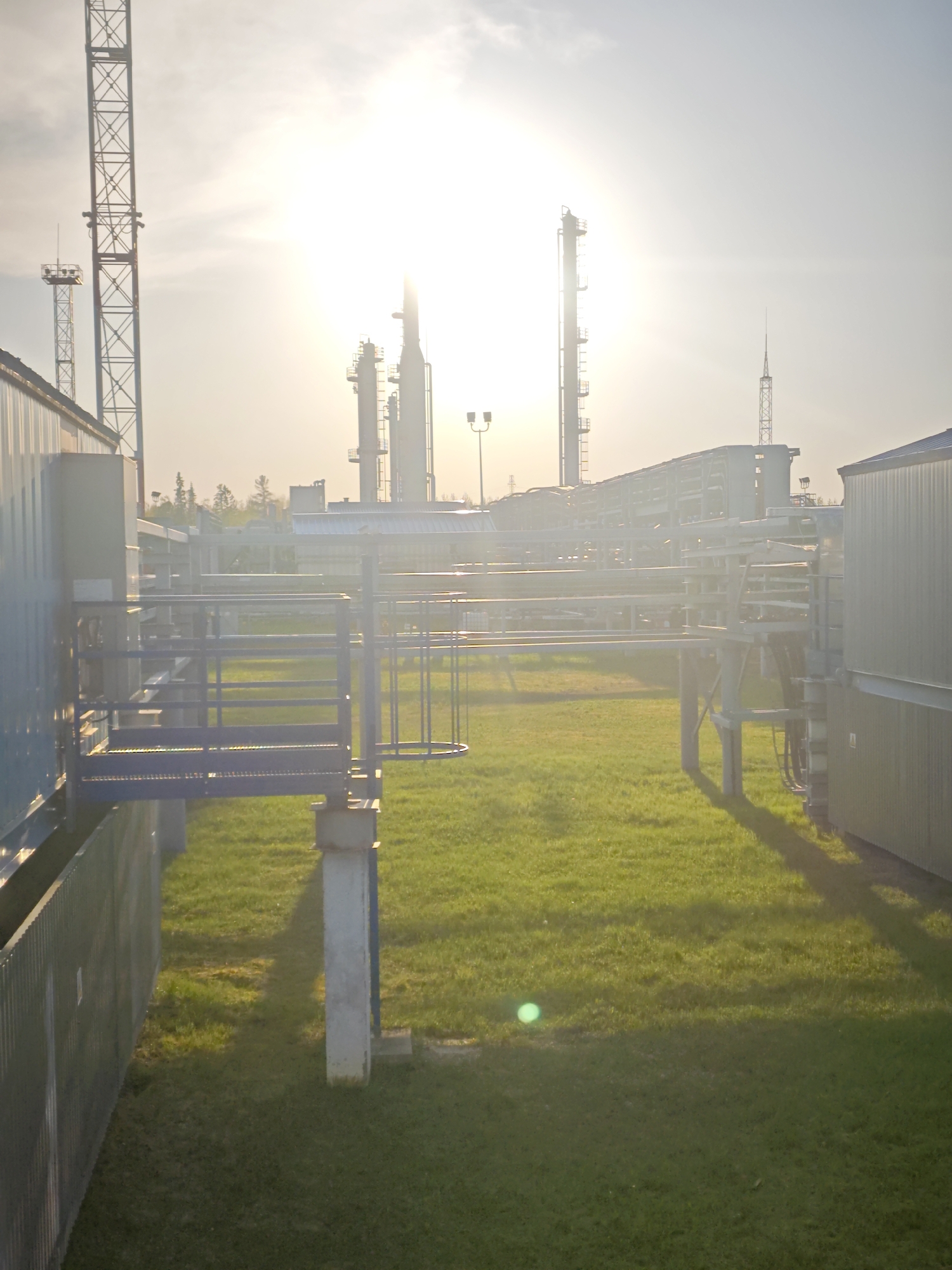The image depicts an industrial facility in Unknown, Russia, dominated by metal structures and bright sunlight. Several towers are visible in the background, suggesting a refinery or processing plant. Above-ground piping spans across a grassy area. A fence is present on the left. Two metal buildings are located on the left and right of the foreground. The sky is obscured by intense sunlight. There are no visible people.