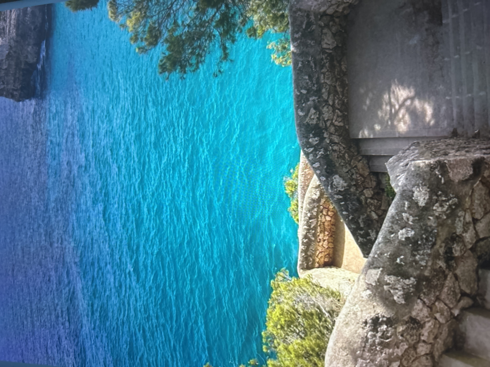 This media file captures a vibrant coastal landscape from an elevated perspective, oriented vertically but depicting a horizontal scene. The dominant feature is a vast expanse of clear water, showcasing a gradient from brilliant turquoise in the foreground to deeper blue further away. The surface of the water is subtly textured with ripples, suggesting calm conditions.
On the left side of the scene, rugged, dark cliffs descend sharply into the water. On the right, a weathered stone railing or balustrade frames the view. This stone structure, appearing to be made of irregular blocks and possibly covered in moss or lichen, forms part of a viewing area or pathway. Green foliage, likely bushes or trees, thrives along the cliff edge and around the stone work, casting soft, dappled shadows onto a paved or stone platform behind the railing. The bright daylight indicates clear weather, possibly late morning or early afternoon, with strong sunlight illuminating the scene. No individuals or specific activities are present, allowing the natural beauty of the rocky coastline and pristine waters to take center stage. There is no visible text in the image. The location cues suggest a Mediterranean or similar coastal region known for its dramatic cliffs and exceptionally clear waters.