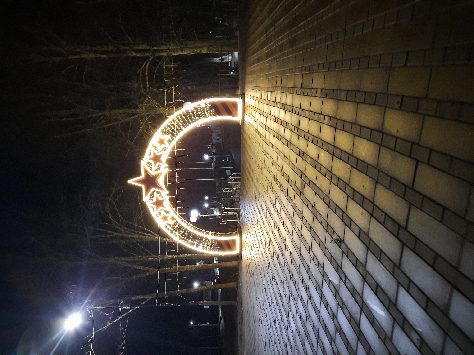 The image depicts a brightly lit decorative structure in Krasnogorskoye, Russia. The structure is a semi-circular arch adorned with stars, illuminated with golden lights, likely for a holiday celebration. It is positioned near a brick wall, which takes up a significant portion of the right side of the image. The scene is captured at night, with bare trees visible in the background. The presence of other streetlights and building lights indicates an urban setting.