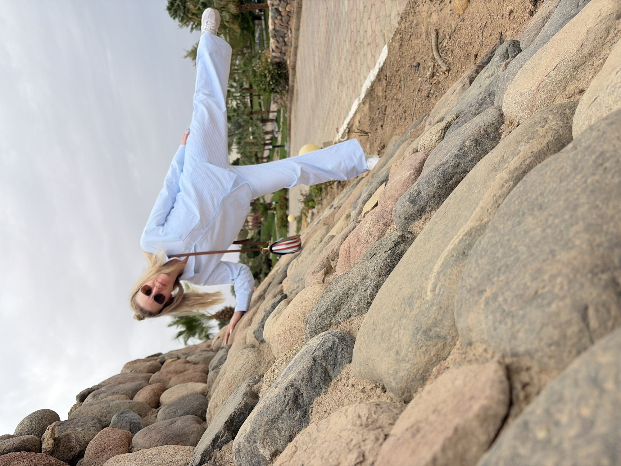 A woman is doing a handstand on a rock wall in Sharm El Sheikh, Egypt. The woman is wearing a light blue striped suit and white sneakers, with sunglasses on her face. She has blonde hair. The rock wall is made of large, rounded stones. Behind the wall, there is a grassy area with trees and a paved path. The sky is overcast. The location is identifiable as Sharm El Sheikh based on the distinctive rock formations and desert landscape.