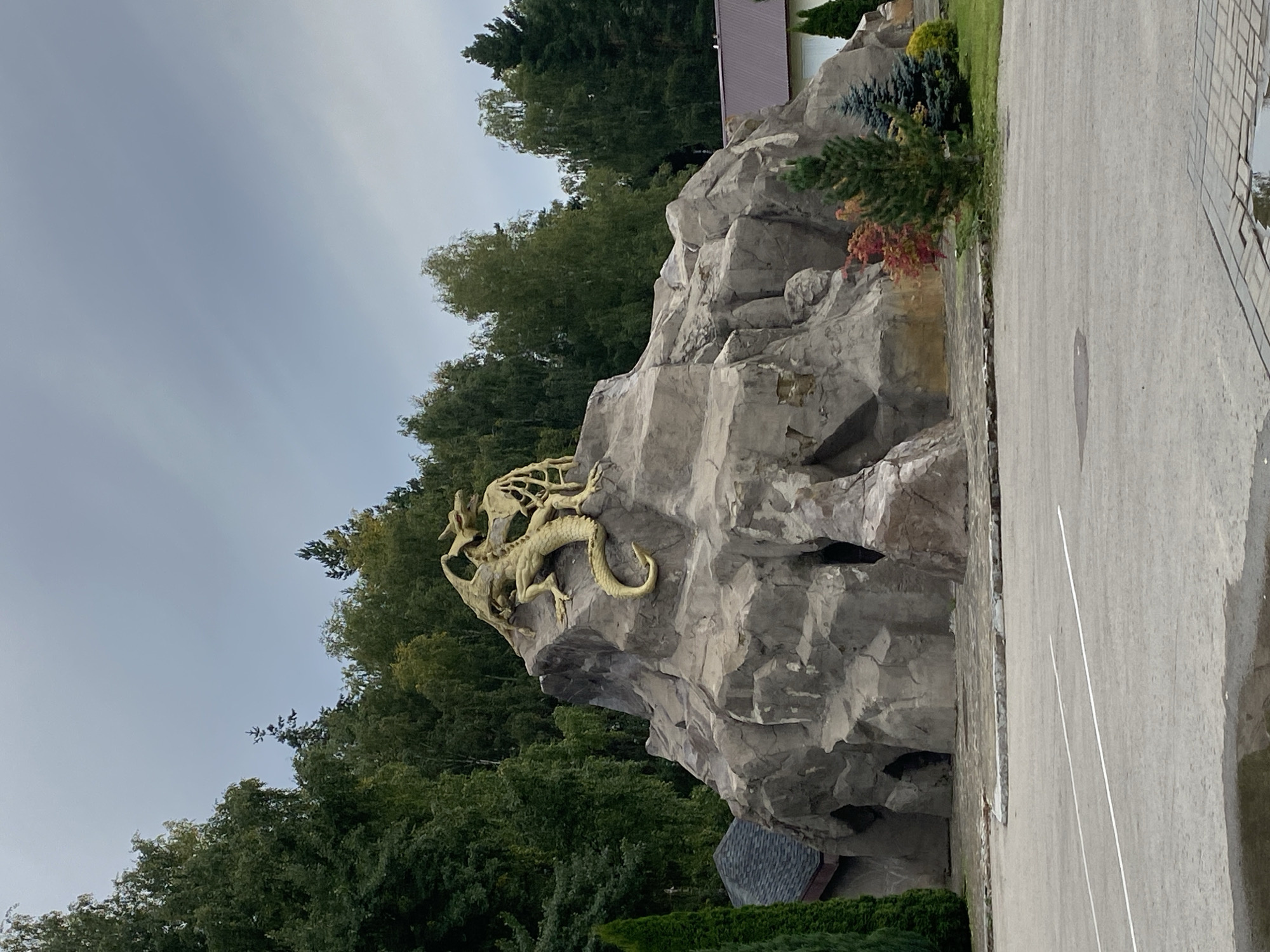 The image shows a large, man-made rock formation topped with a golden statue of a dragon. The rock formation has several openings that resemble caves. The dragon statue is detailed, with wings, claws, and a long tail. The rock formation is situated in an outdoor area with trees in the background and a paved area in the foreground, which appears to be a parking lot or plaza. There are buildings visible to the right and left of the rock formation. The sky is overcast. The location is identified as being in Chulis, Russia. There are no people visible in the image.