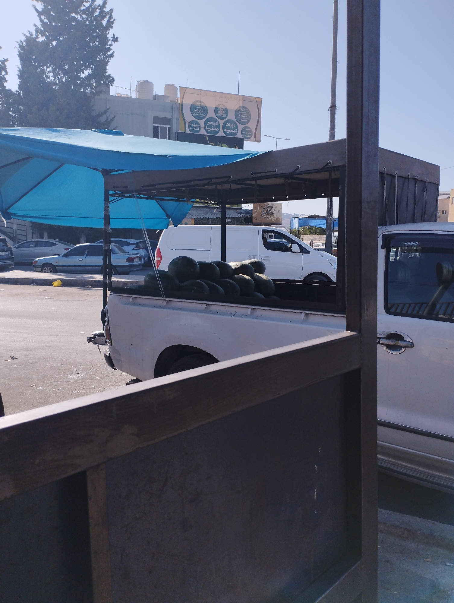 An outdoor marketplace scene is depicted in Baqa'a camp, Jordan, under a clear blue sky. A white pickup truck is parked, its bed filled with a large quantity of watermelons. The truck is partially covered by a dark fabric awning supported by metal poles, and in front of this, a bright blue tarp extends over a frame. In the background, several cars are visible on a paved street, and a white van is parked. Buildings and trees form the backdrop, with a billboard featuring Arabic text. The foreground is dominated by a dark, metallic structure, possibly a railing or part of a stall, obscuring a portion of the view. The overall impression is of a vendor's stall in a bustling urban environment.