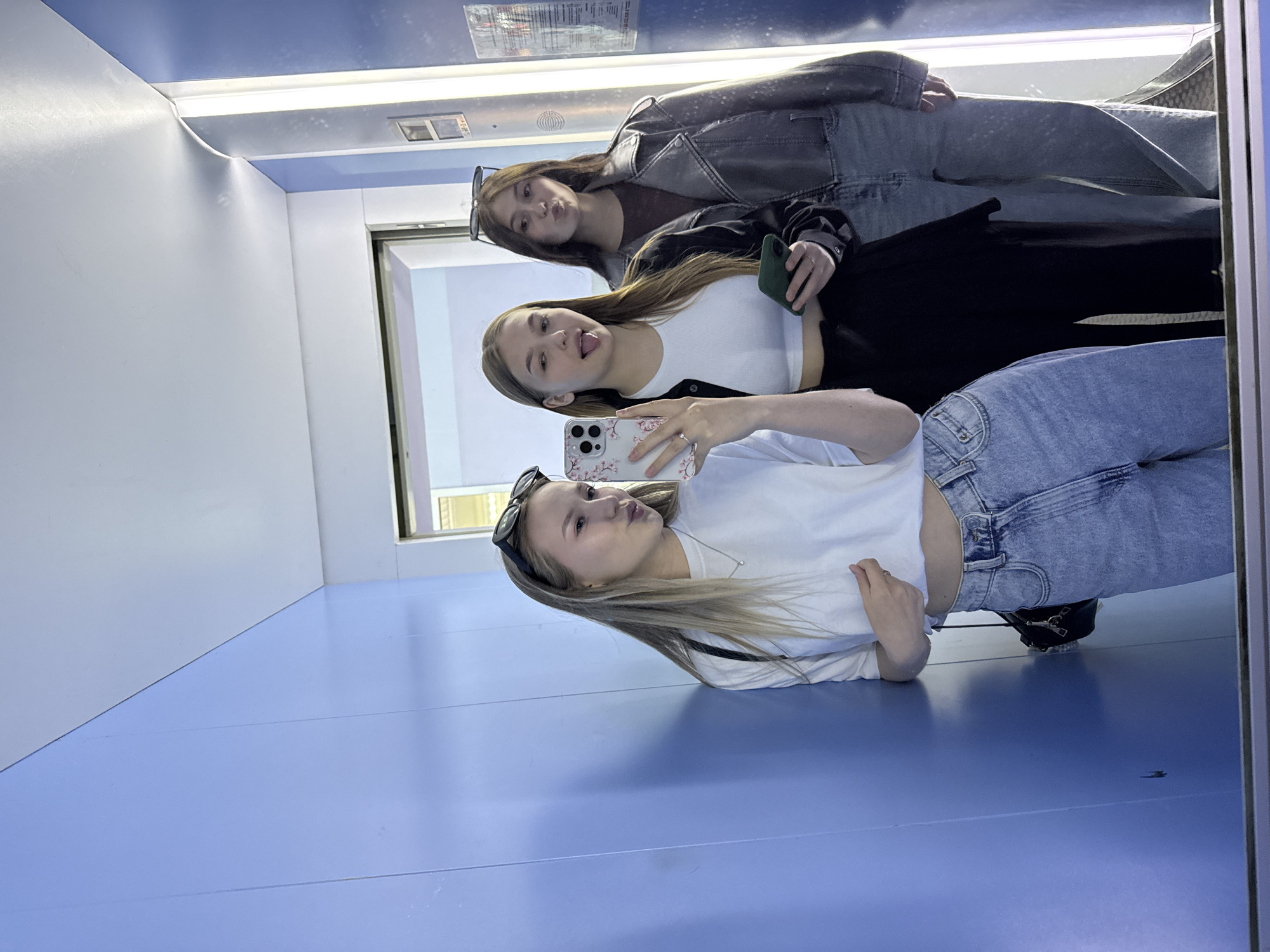 Three young women are taking a mirror selfie in what appears to be an elevator or a narrow corridor with blue walls. The woman on the left is wearing a white t-shirt, light-wash jeans, and sunglasses perched on her head. She is holding a phone and a black purse. The woman in the middle is wearing a white crop top, black pants, and is holding a green phone and her phone with a floral case. She is sticking her tongue out. The woman on the right is wearing a dark brown shirt and a grey leather jacket with light-wash jeans, and sunglasses on her head. She is puckering her lips. The environment is brightly lit, with a vertical light fixture visible on the right. The overall scene suggests a casual and playful moment captured by the individuals. The location is identified as Chaikovsky, Russia.