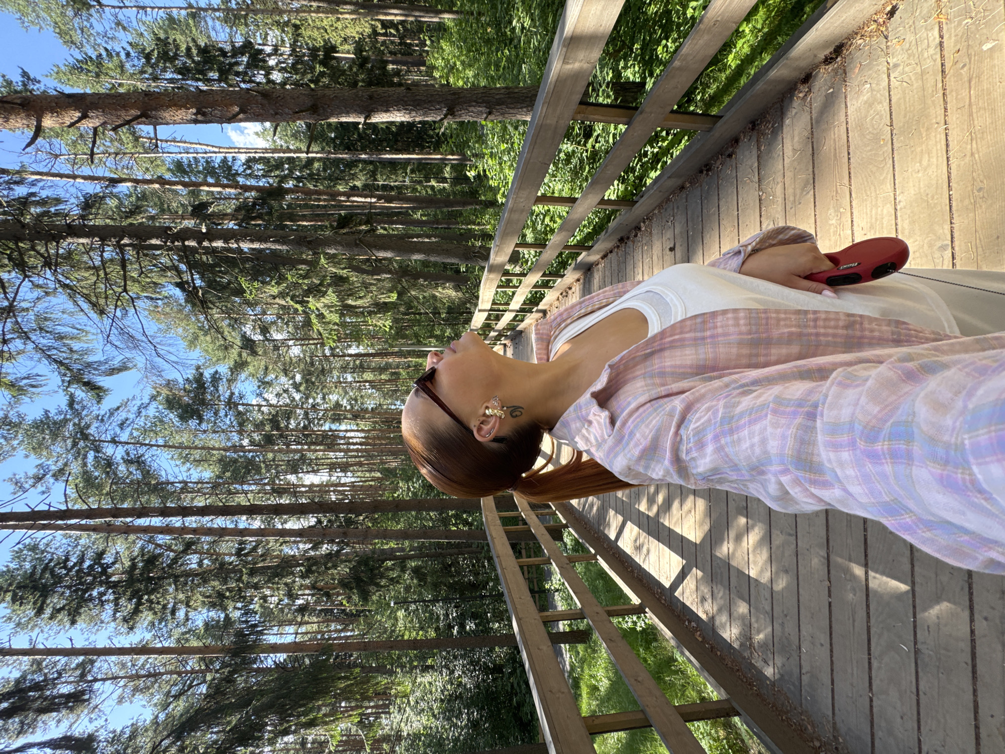 A young woman is lying on a wooden walkway in a forest. She is wearing a white tank top and a pastel plaid shirt. Her head is tilted back, and she is looking up at the sky. A red retractable dog leash is in her hand. The sun is shining through the trees, casting dappled shadows on the walkway. The forest appears to be made up of tall pine trees. The time of day appears to be late afternoon, given the long shadows. The weather is sunny and warm. The location is Votkinsk, Russia. There is no visible text in the image.