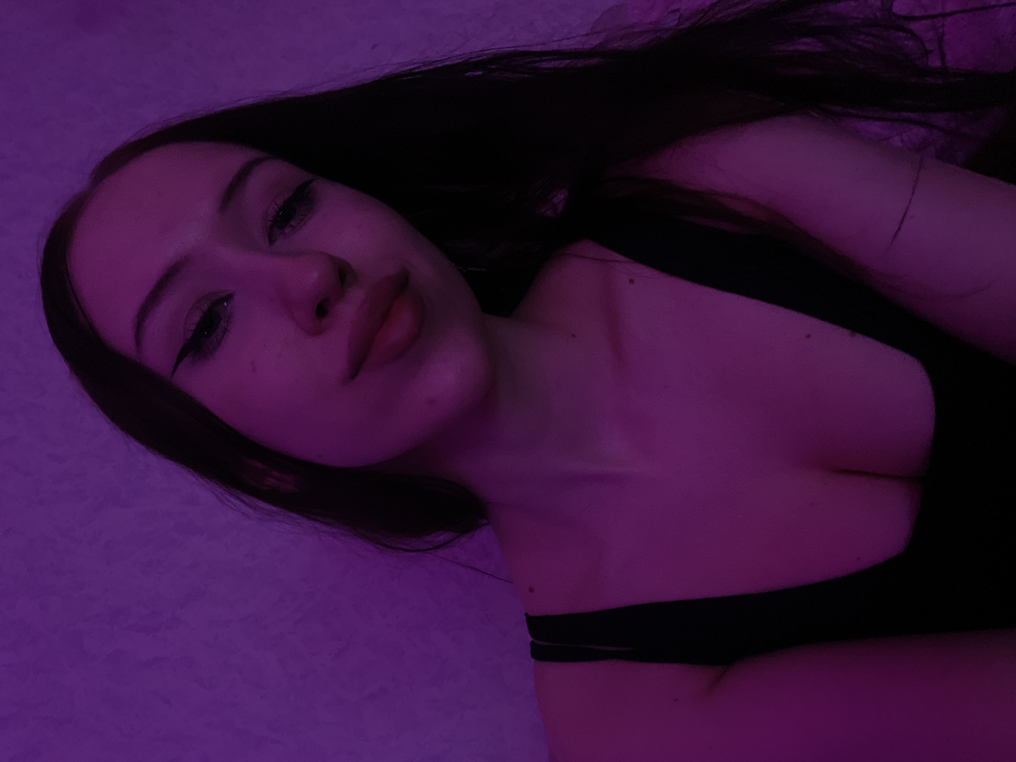 A young woman with long, dark hair is shown in a close-up, selfie-style portrait. She is wearing a black top, and her makeup includes winged eyeliner. The lighting in the image is predominantly purple, casting a distinct hue over her skin and the textured background, which appears to be a wall. The scene is indoors, and no specific geographical landmarks of Votkinsk, Russia, are visible or identifiable.