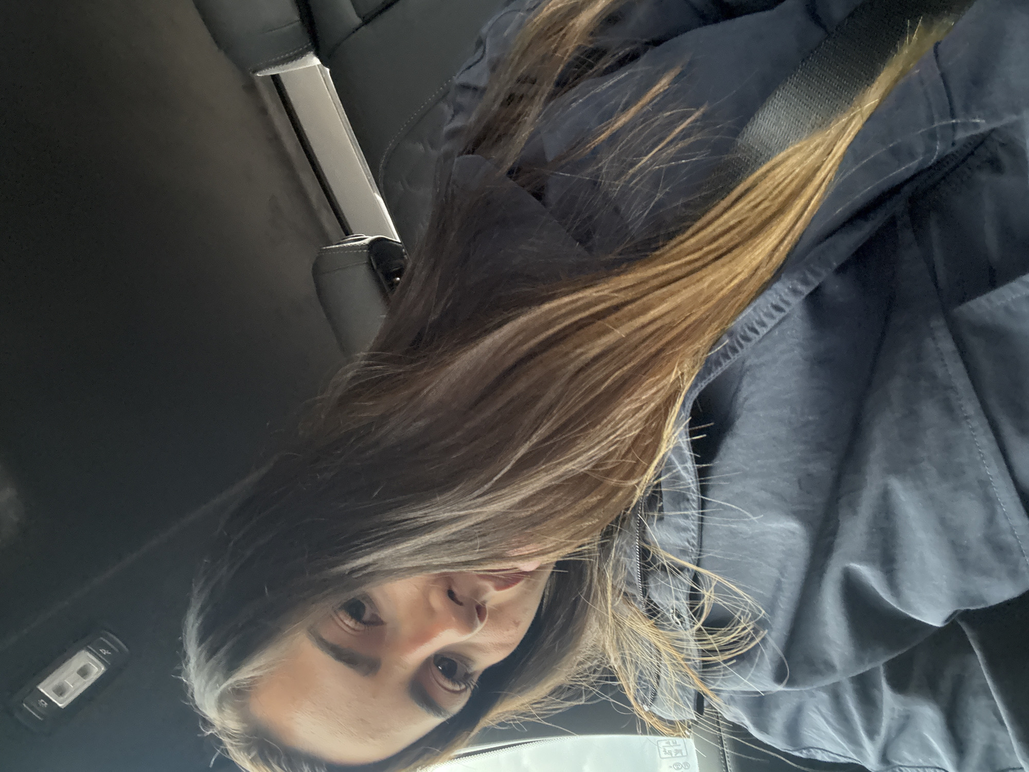 A woman is seated in a car, wearing a seatbelt and a blue jacket. Her hair is long and brown. The location is not discernible from the image; Santa Rita Tlahuapan, Mexico cannot be confirmed. The interior of the car is visible, including a headrest, seat controls, and the ceiling.