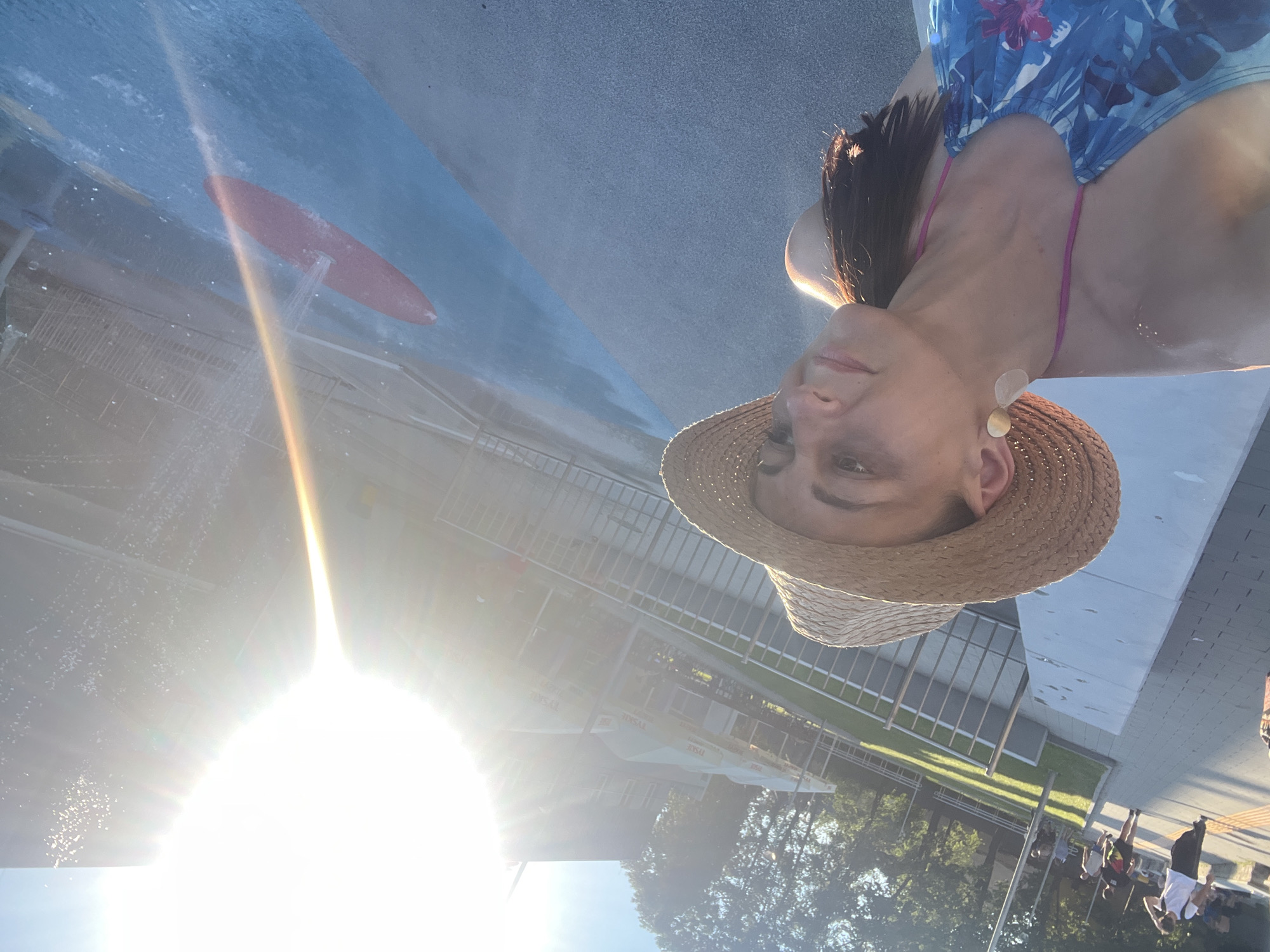 A woman wearing a straw hat and a blue and pink floral swimsuit is taking a selfie in an outdoor splash pad area. The sun is bright and creating lens flare. In the background, there are trees, buildings with umbrellas in front, and a few people walking on a paved path. The splash pad has blue and grey surfaces, with a red oval shape visible in the blue section, and water spraying from an unseen source.