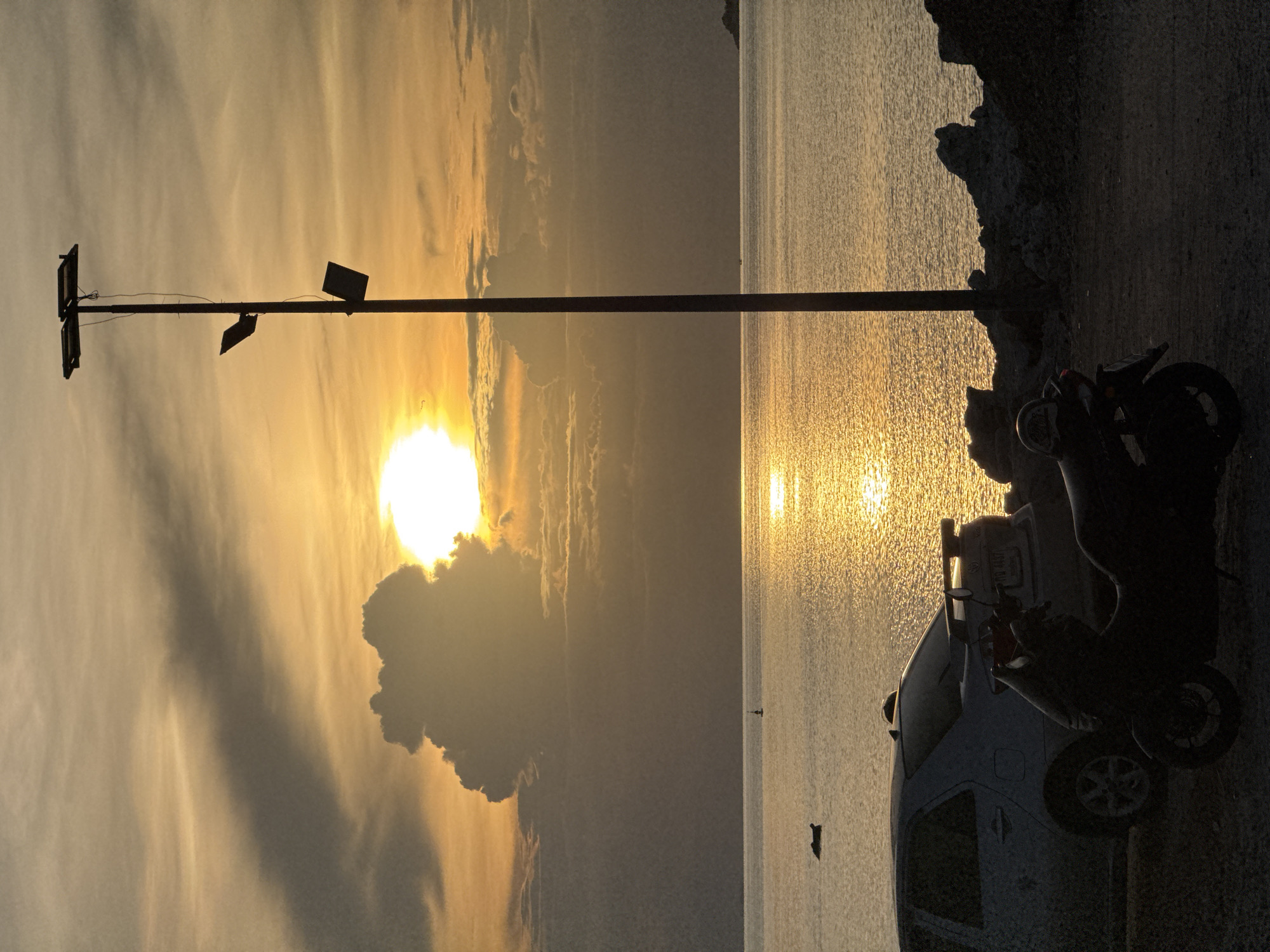 The image depicts a sunset scene by the sea. The sun is low on the horizon, casting a warm, golden glow across the water and the sky, which is filled with dramatic clouds.
The subjects include a street light with two fixtures, a parked car, and a scooter. The car is light blue and its rear is facing the viewer, while the scooter is parked in front of it and is silhouetted against the sunset. There are also two small, dark figures visible in the water, possibly swimmers or small boats. The scene is set along a rocky coastline with a paved area where the vehicles are parked.
The time of day is clearly sunset, given the low angle of the sun and the warm, diffused light. The weather appears to be clear with some cloud cover. The location is identified as Koh Phangan, Thailand. The overall emotion evoked is one of tranquility and the beauty of nature.
There is a license plate visible on the back of the car, but the text is illegible.