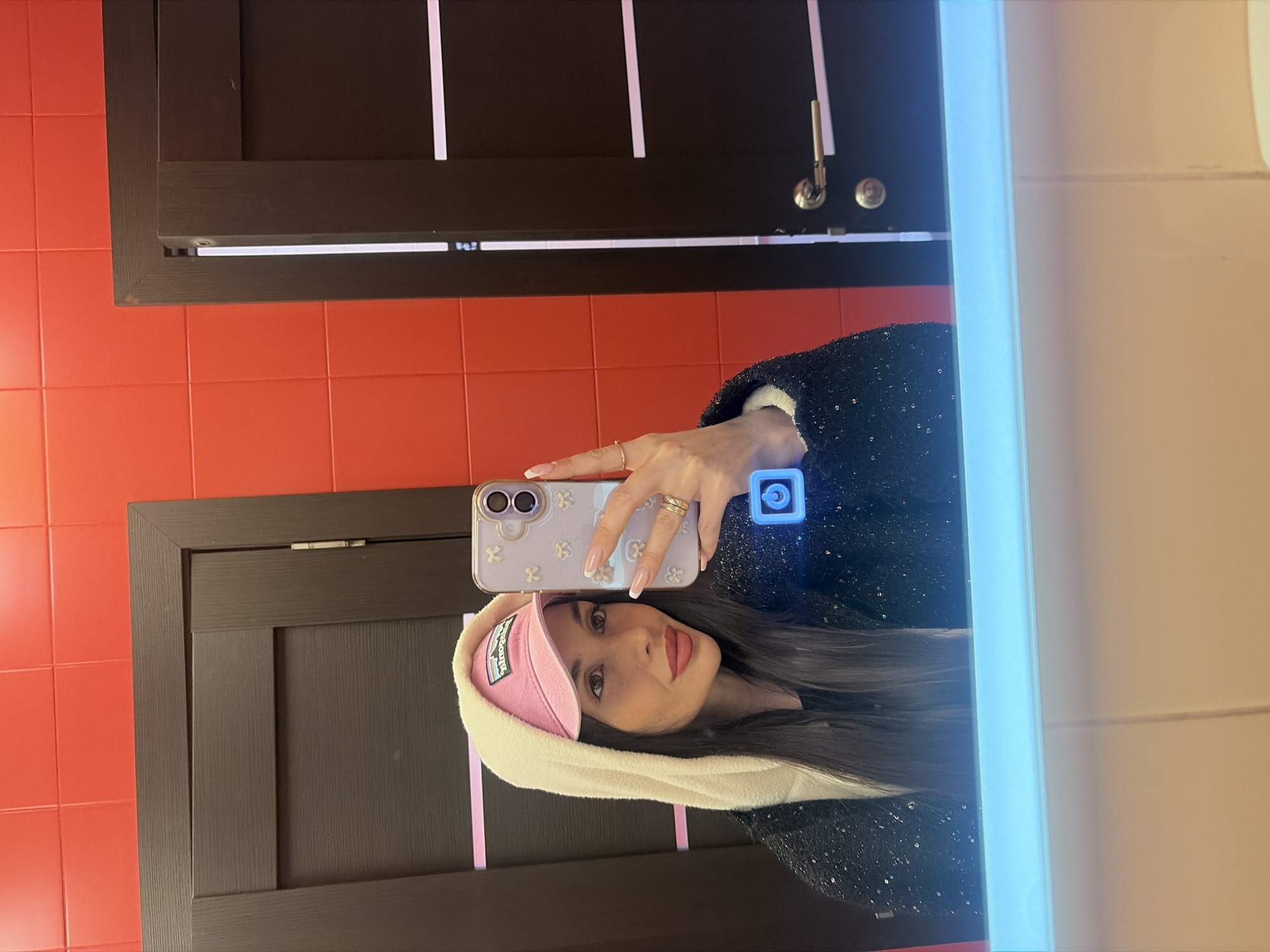 A woman is taking a selfie in a bathroom. The woman is wearing a black sequined jacket and a pink hat with a white lining. She is holding a white smartphone in her left hand with a visible case, and her fingers are adorned with rings. The background includes orange tiles, a dark wooden cabinet with light-colored vertical stripes, and a mirror with a light. The location appears to be a bathroom, but there is no specific information to determine if it is located in the city of Saint Petersburg, Russia.